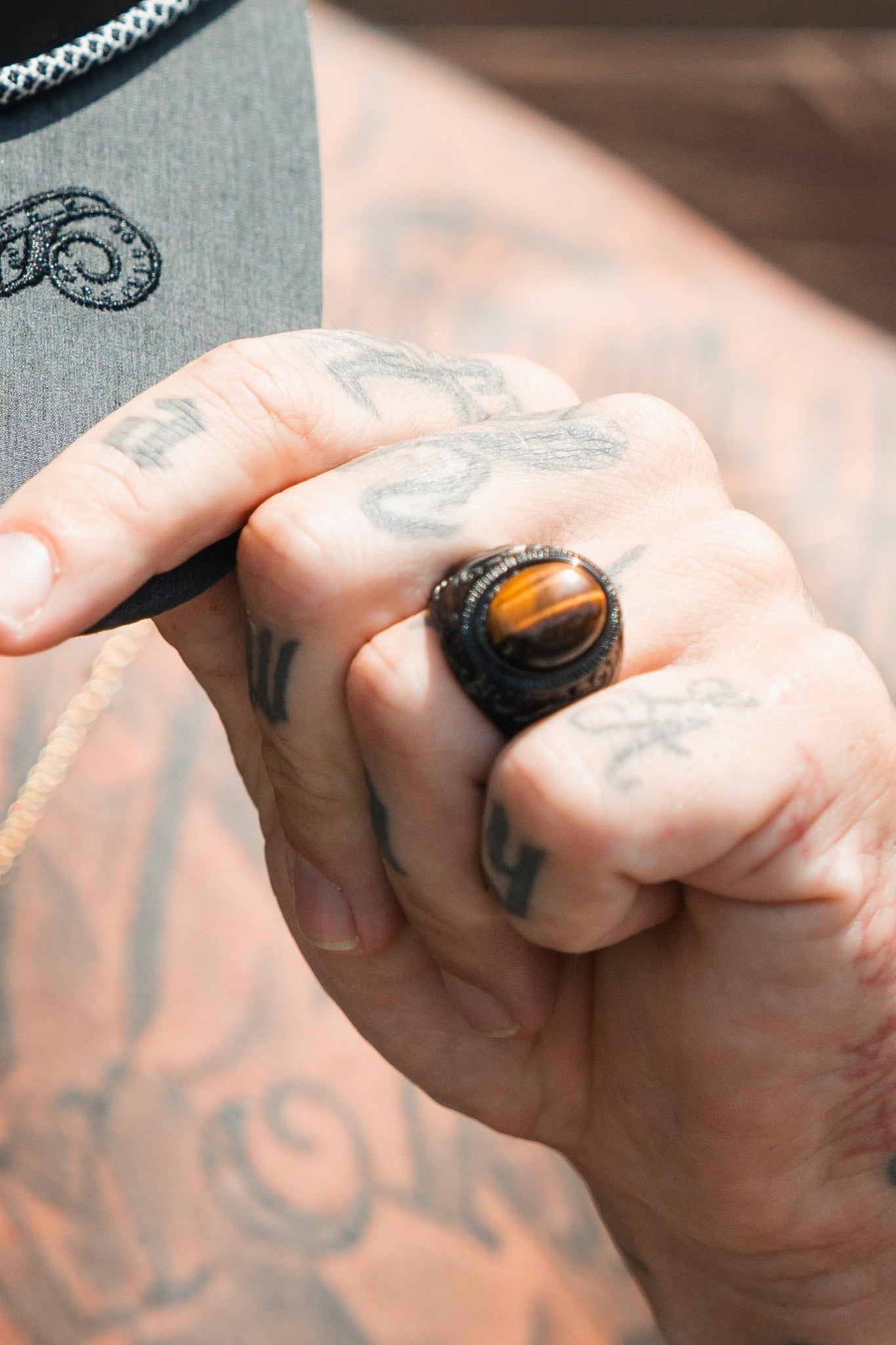 A close-up shows tattooed fingers holding a hat brim, featuring the Eye Of The Tiger Ring by Relentless Betrayal. The tanned skin displays letters and designs, enhanced by the ring’s unique flair against a softly blurred background.