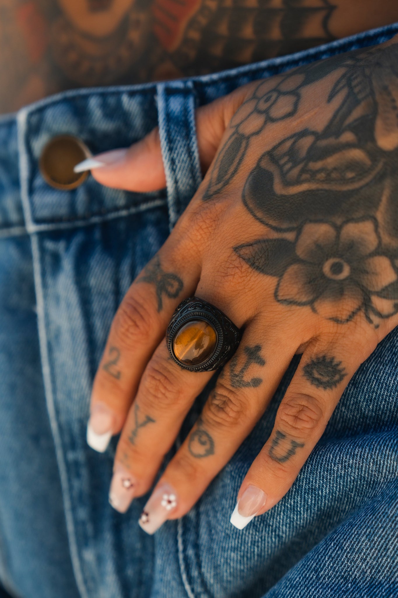 A close-up reveals a tattooed hand on a denim waistband, adorned with Relentless Betrayals Eye Of The Tiger Ring. The hand also features detailed nail art, with clear polish and decorative elements, showcasing unique style and personality in handcrafted jewelry.