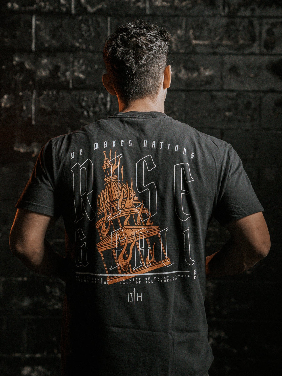 A person stands facing a dark, textured wall, wearing the Rise & Fall Premium Tee by 13th. The black T-shirt features an orange ship graphic and the words He Makes Nations Rise & Fall on the back. The dim lighting creates a dramatic mood, capturing the essence of lifes fleeting moments.