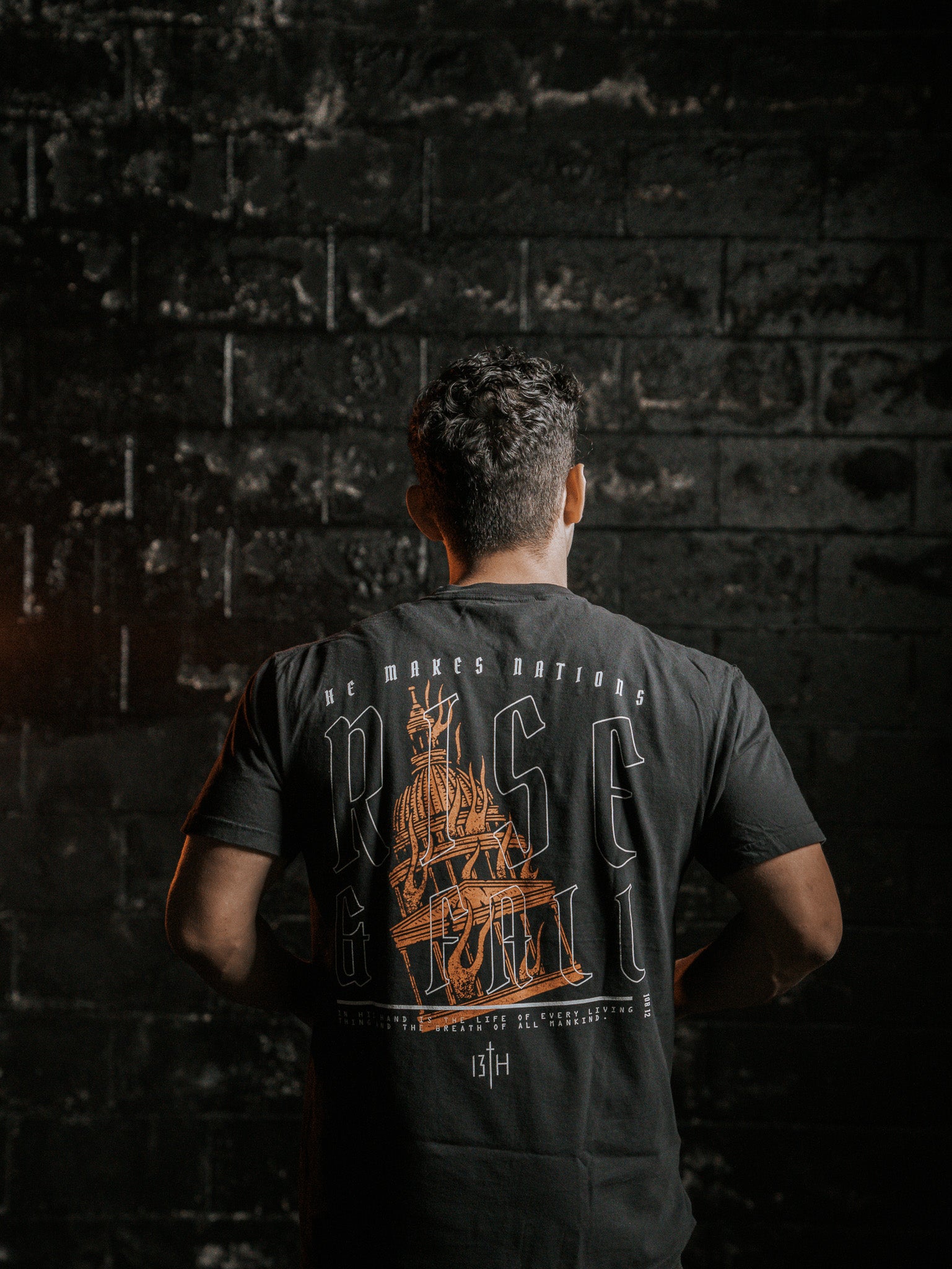 A person stands facing a dark, textured wall, wearing the Rise & Fall Premium Tee by 13th. The black T-shirt features an orange ship graphic and the words He Makes Nations Rise & Fall on the back. The dim lighting creates a dramatic mood, capturing the essence of lifes fleeting moments.
