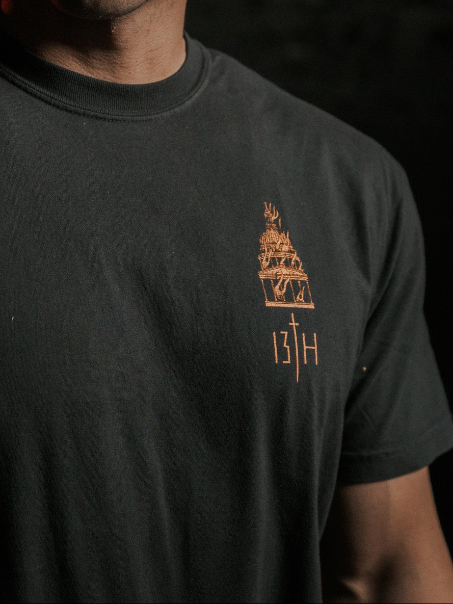 A man with short hair stands in a dimly lit room, embodying lifes essence. He wears the Rise & Fall Premium Tee by 13th, featuring a subtle design and the number 13H, symbolizing humanitys mysterious journey amidst a dark, shadowed background.