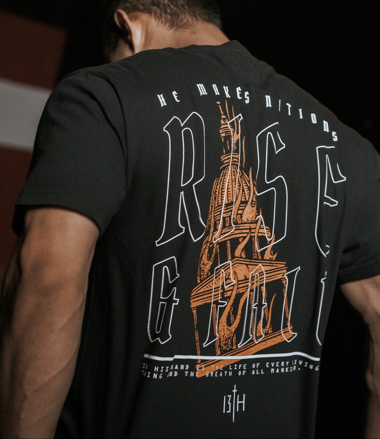 A person with short dark hair is seen from behind, wearing the black Rise & Fall Premium Tee by 13th. The T-shirt features large text RISE, a stylized building illustration, and small decorative text symbolizing mankinds journey.