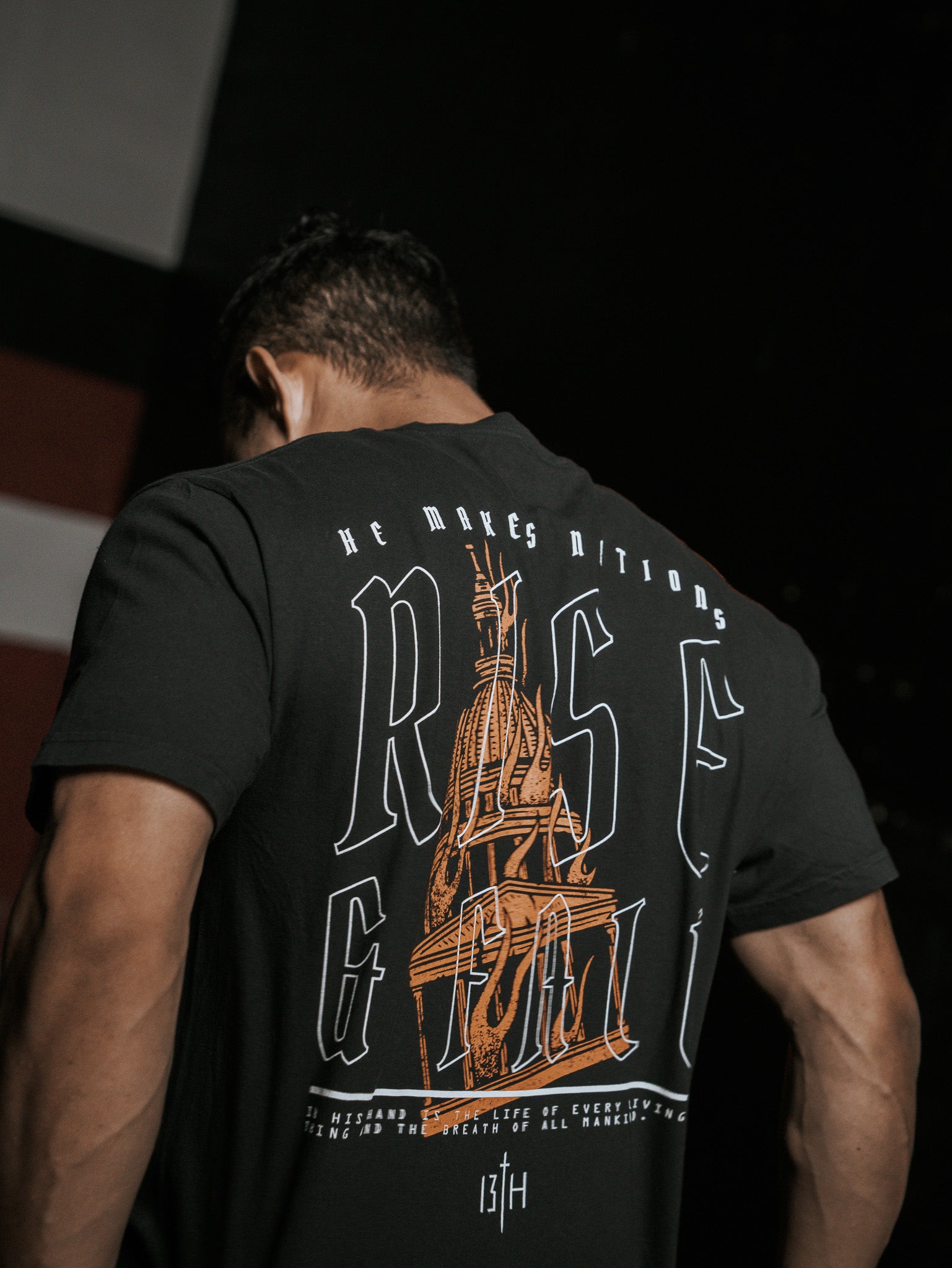 A person with short dark hair is seen from behind, wearing the black Rise & Fall Premium Tee by 13th. The T-shirt features large text RISE, a stylized building illustration, and small decorative text symbolizing mankinds journey.