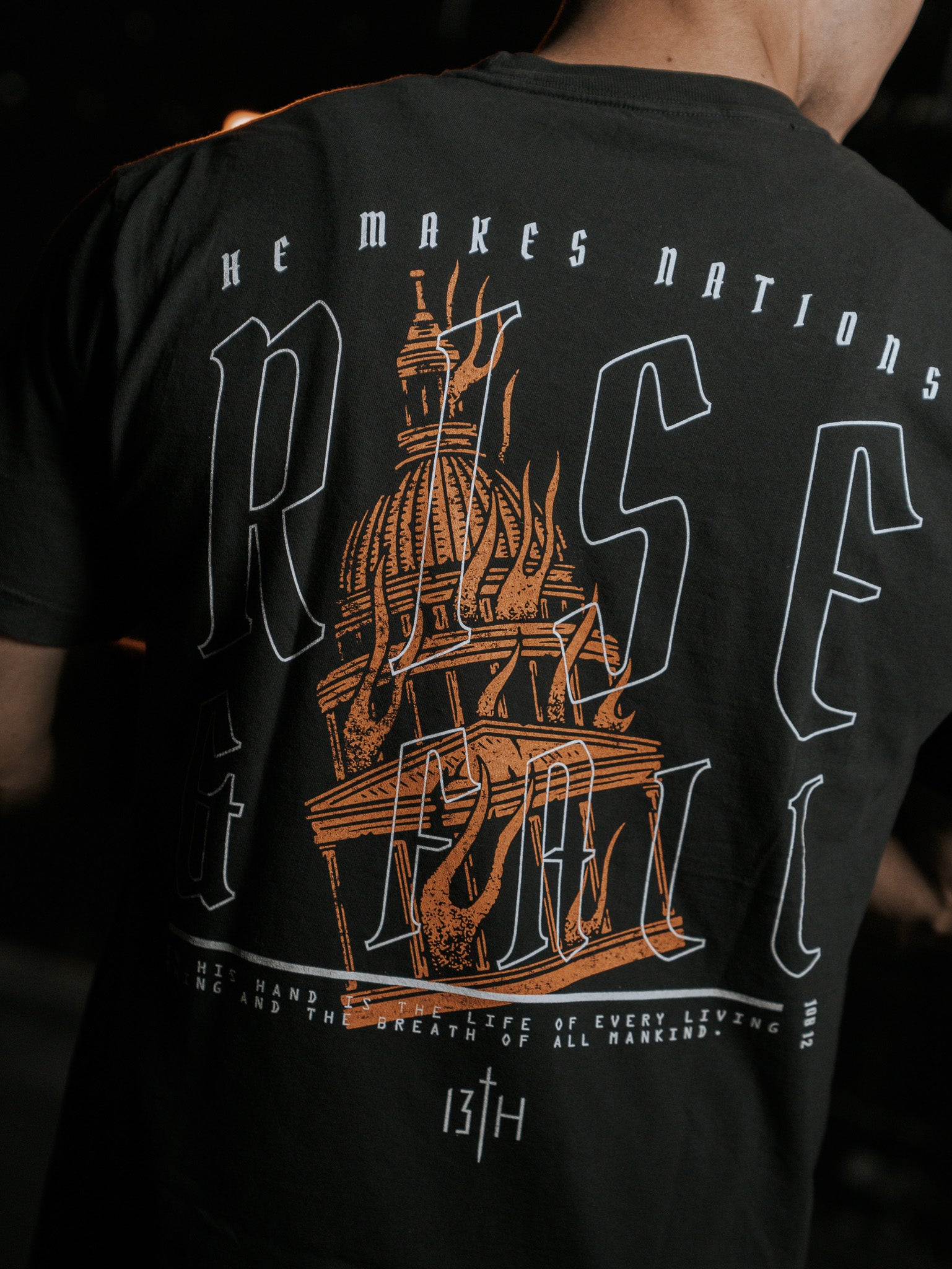 Back view of a person in the black Rise & Fall Premium Tee by 13th, featuring a burning candle inside a dome and the text He makes nations rise & fall and The life of every living thing and the breath of all mankind.