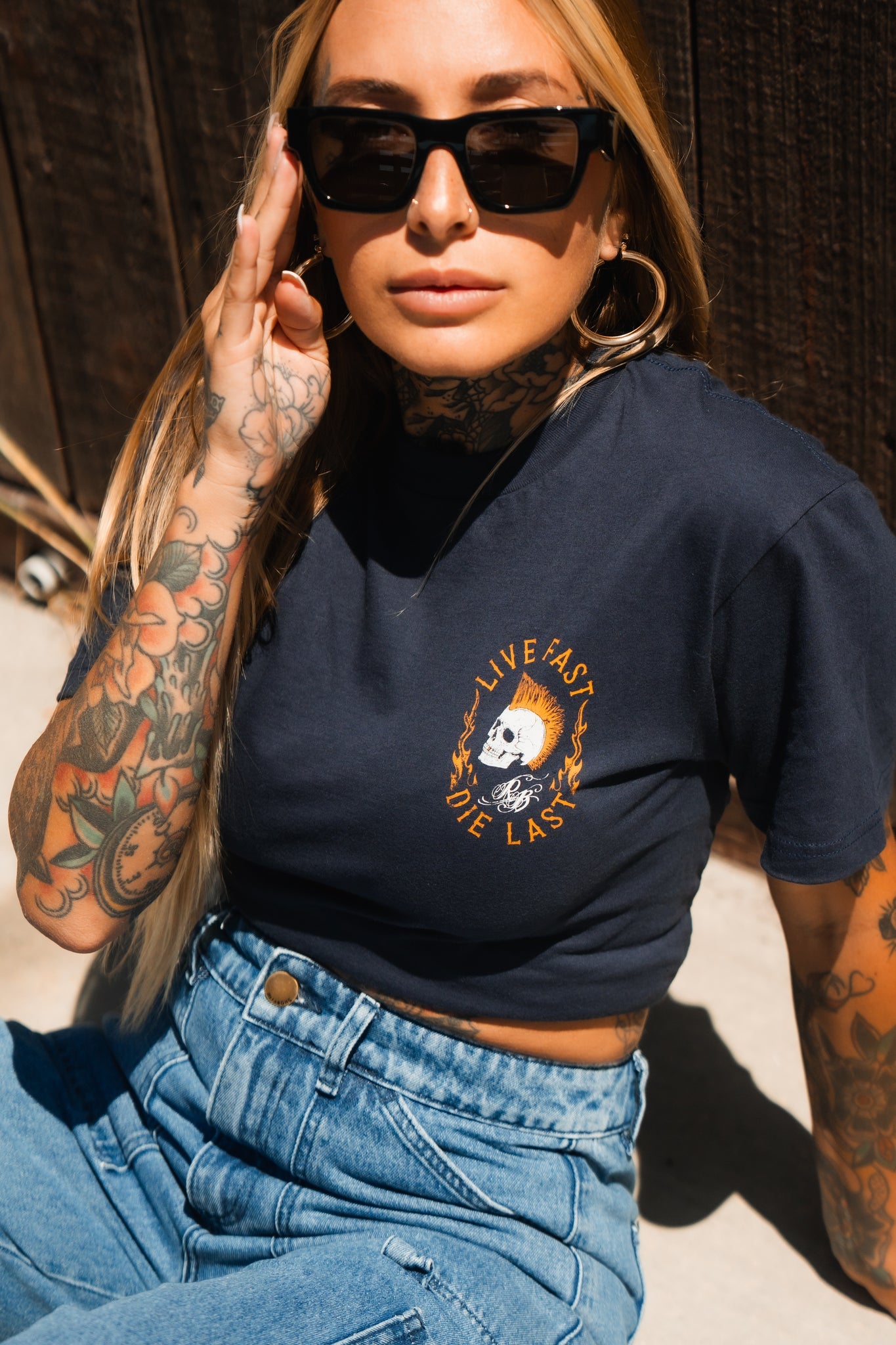 A person embodying youth culture and adventure sits outdoors with long hair, tattoos, and sunglasses. Theyre wearing Relentless Betrayals Live Fast Navy T-Shirt and high-waisted jeans. Posing confidently, one hand casually touches their sunglasses.