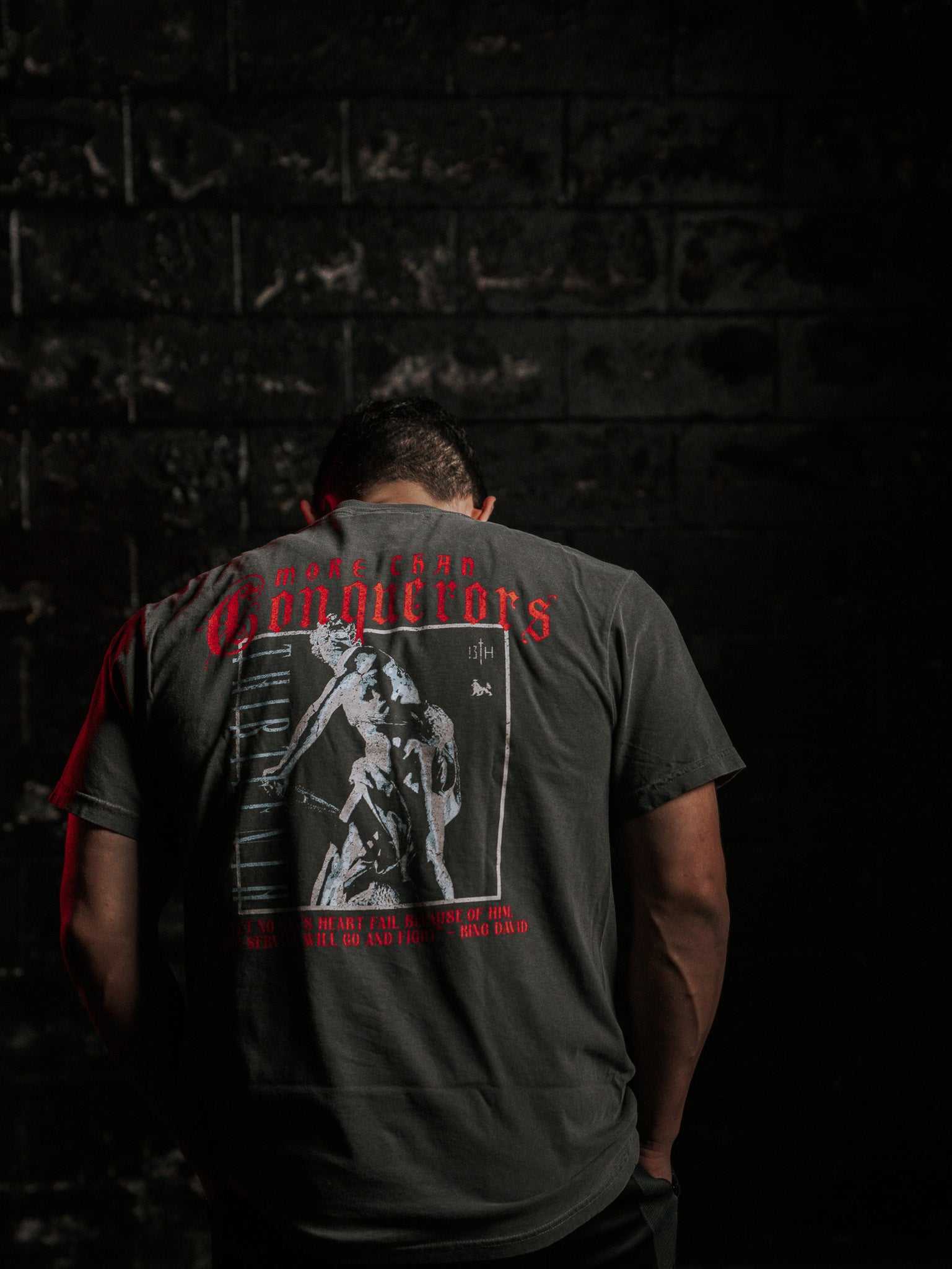 A person stands with their back to the camera, wearing the 13th brand’s Conquerors Premium Tee featuring a graphic that partially reads King David. The dark and moody background, highlighted by red lighting, creates an atmosphere of a valiant fight.