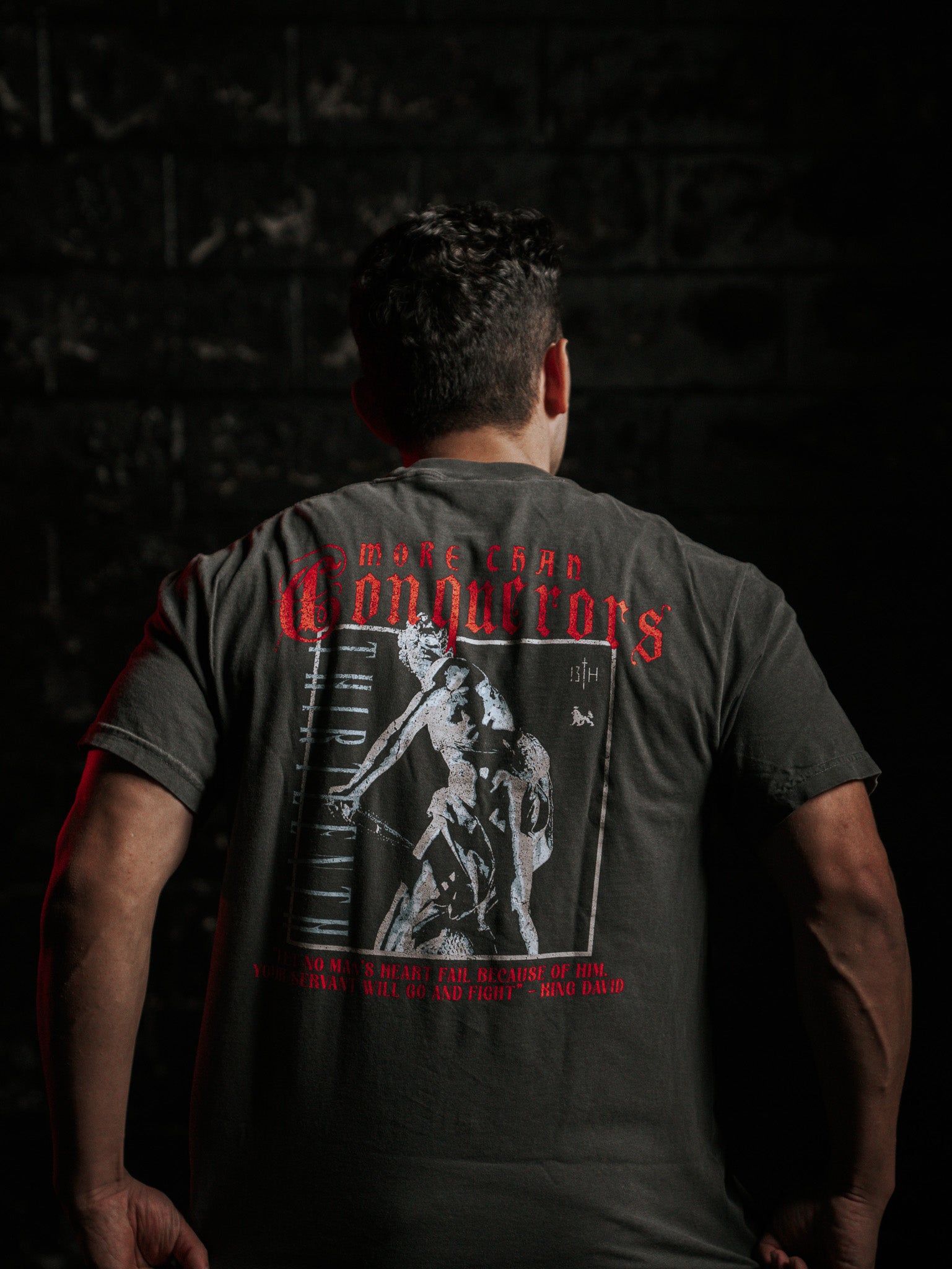 A person is shown from behind, wearing a Conquerors Premium Tee by 13th, featuring red MORE CHAD CONQUERORS text and King David warrior art. The dark backdrop subtly spotlights their determined stance.
