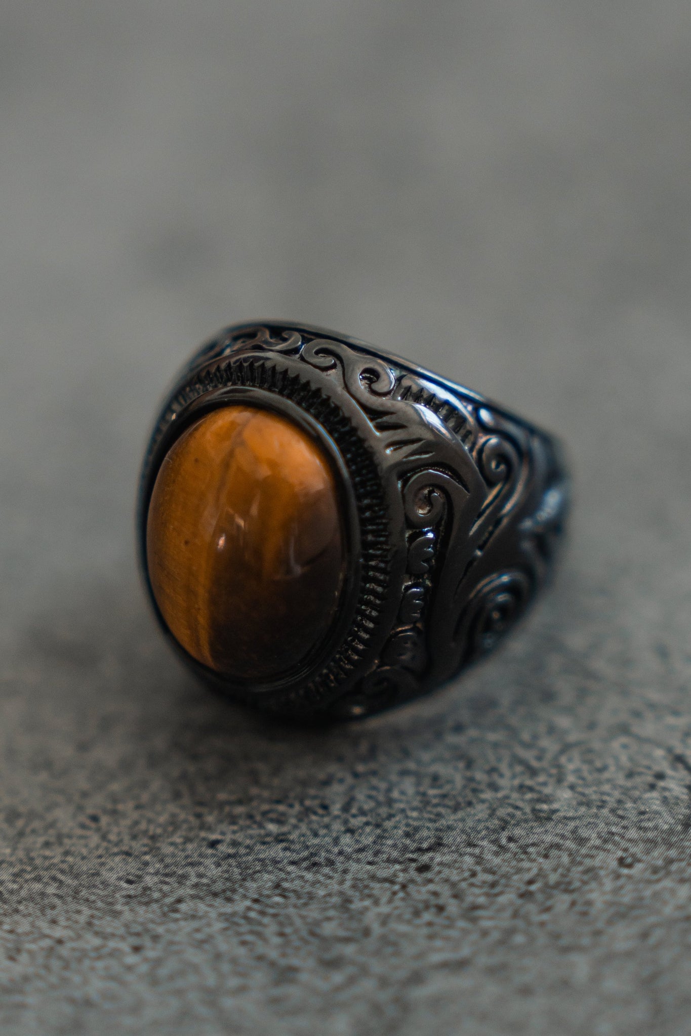 The Eye Of The Tiger Ring by Relentless Betrayal features a detailed stainless steel design, ornate engravings, and an oval tiger eye gemstone with golden-brown bands, set against a textured gray surface.