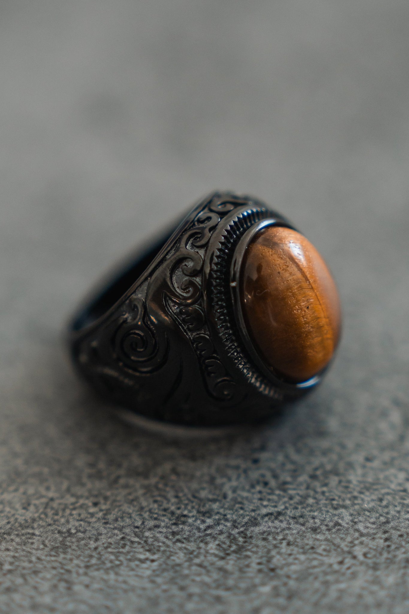 The Eye Of The Tiger Ring by Relentless Betrayal showcases a polished oval tiger eye stone set in a dark, intricately carved setting and displayed on a textured gray surface.