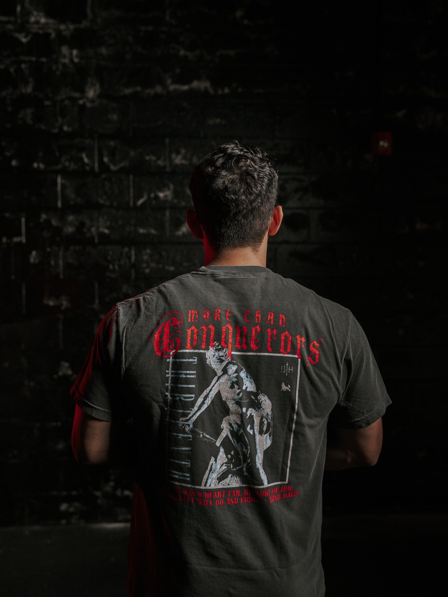 Against a dark, textured wall, someone wears the Conquerors Premium Tee by 13th, adorned with More Than Conquerors in red. Inspired by King Davids spirit, it depicts an athletic figure. Soft lighting highlights the image and words elegantly.