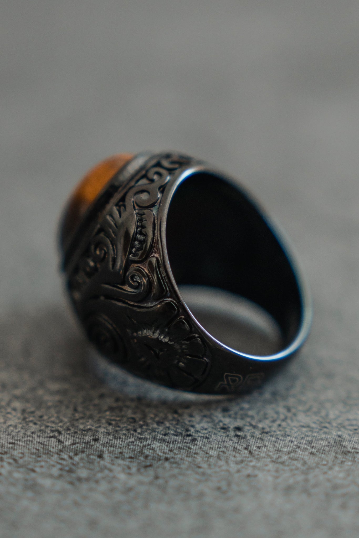The Eye Of The Tiger Ring by Relentless Betrayal, a handcrafted silver piece with intricate engravings and a polished amber stone on top, captivates when displayed on a textured gray surface.