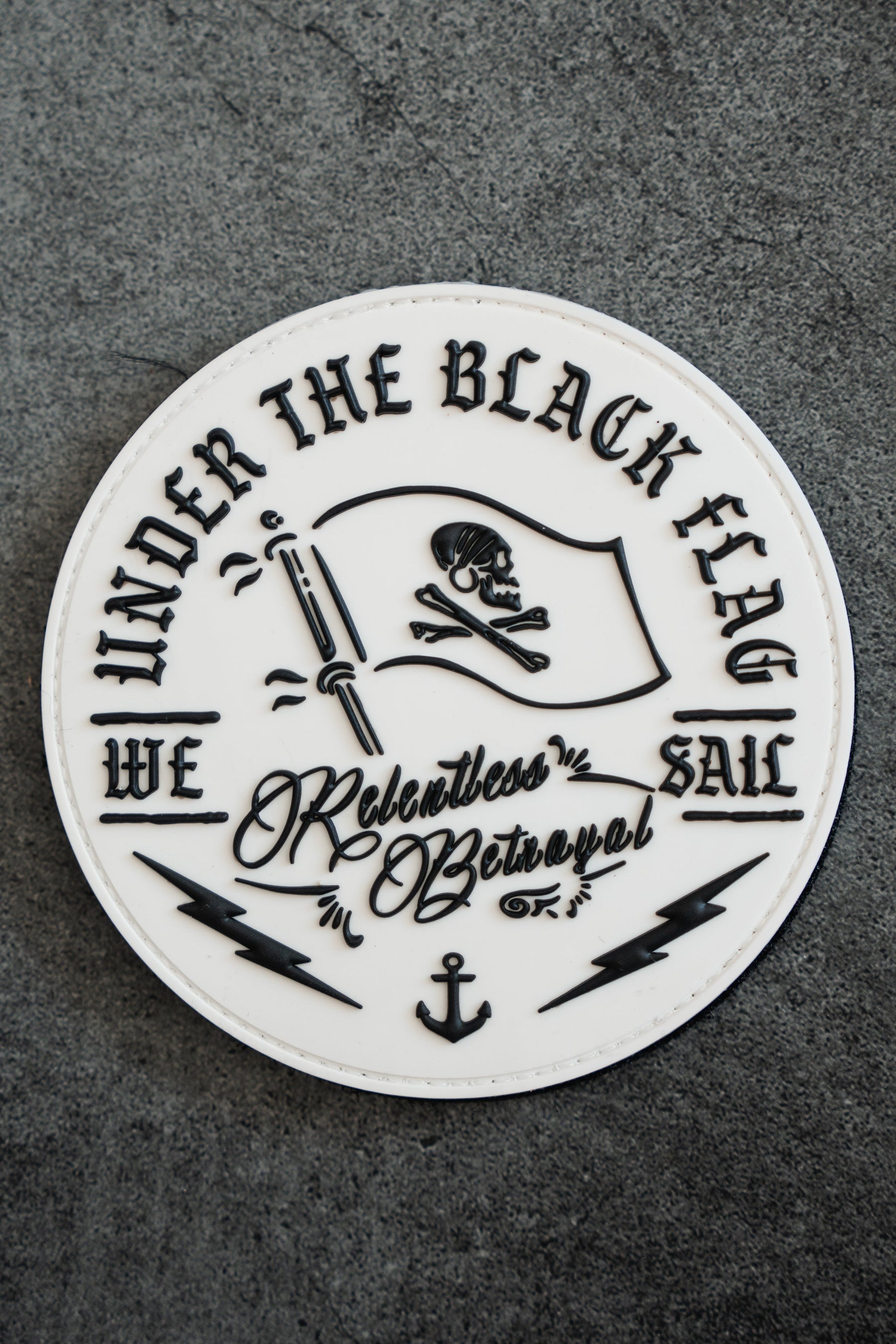 The Black Flag PVC Patch by Relentless Betrayal features a skull and crossbones on a black flag with the text Under the Black Flag We Sail. Set against a textured gray background, it includes lightning bolts and an anchor design.