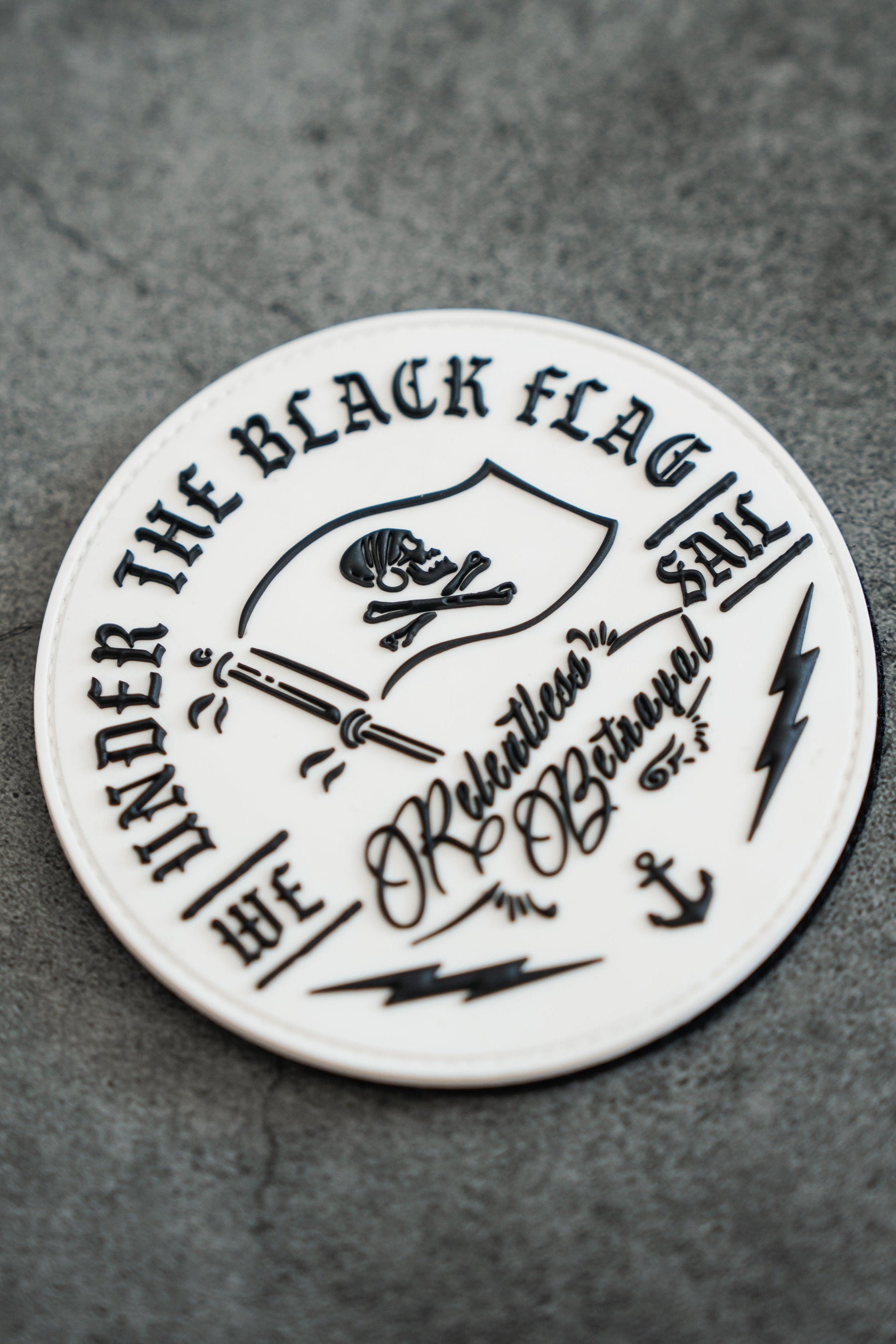 The Black Flag PVC Patch from Relentless Betrayal features a black skull and crossbones at its center with bold text reading, Under the Pirates Black Flag, We Sail, along with the words Rebellious and Obstinate. It is adorned with striking lightning bolt and anchor illustrations.