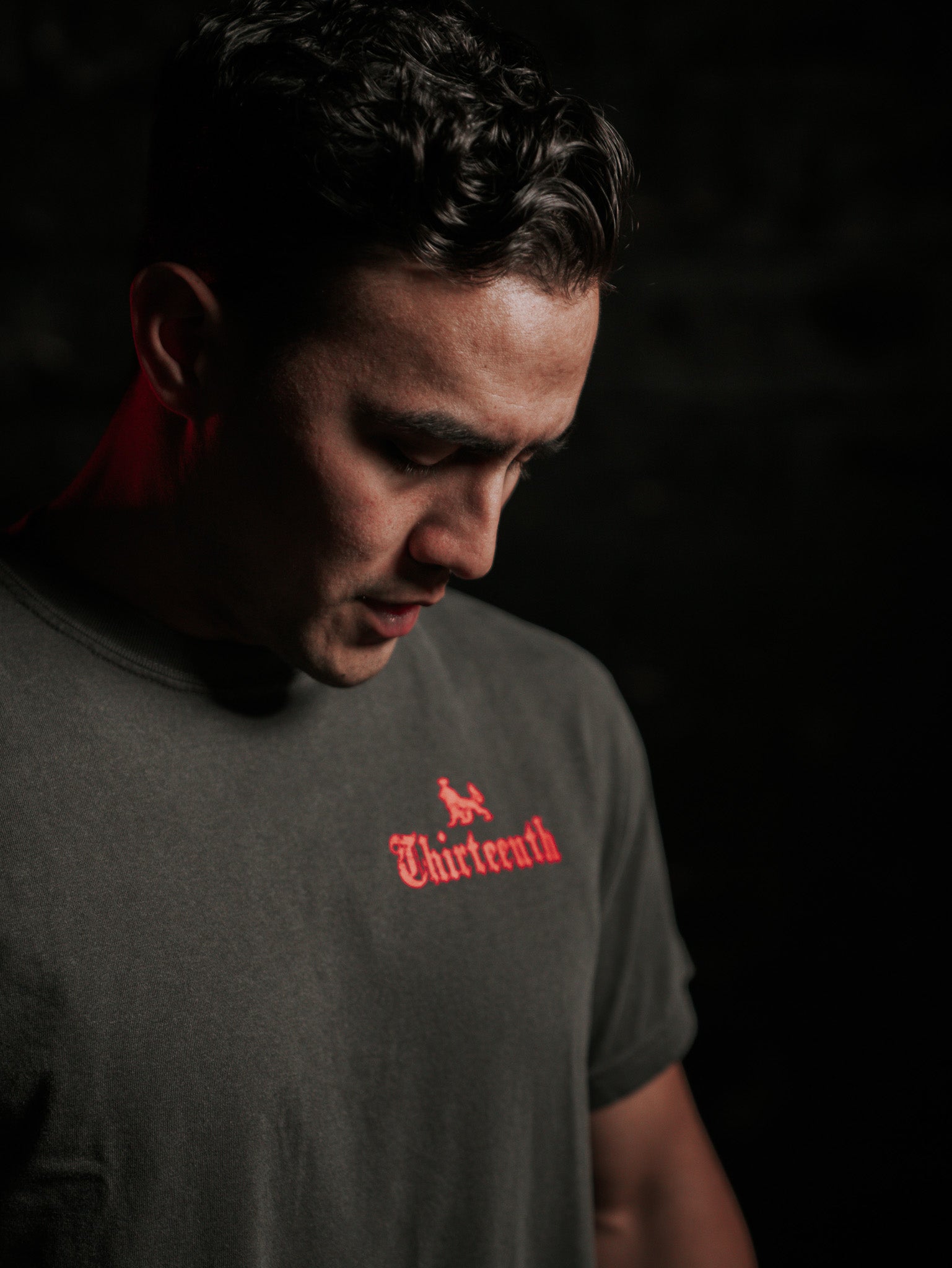 A person with short dark hair looks downward against a dark background, reminiscent of No Mans Heart Fail. They wear the Conquerors Premium Tee by 13th, featuring Thirteenth in red. Subtle red light accents their profile, adding drama to the scene.
