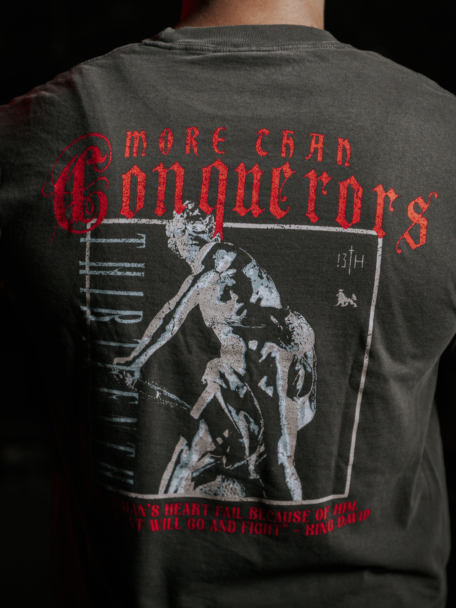 The image shows the back of a person wearing the Conquerors Premium Tee by 13th. The dark t-shirt has a warrior graphic and More Than Conquerors in red, inspired by King Davids legacy, with No Mans Heart Fail boldly printed to evoke bravery.