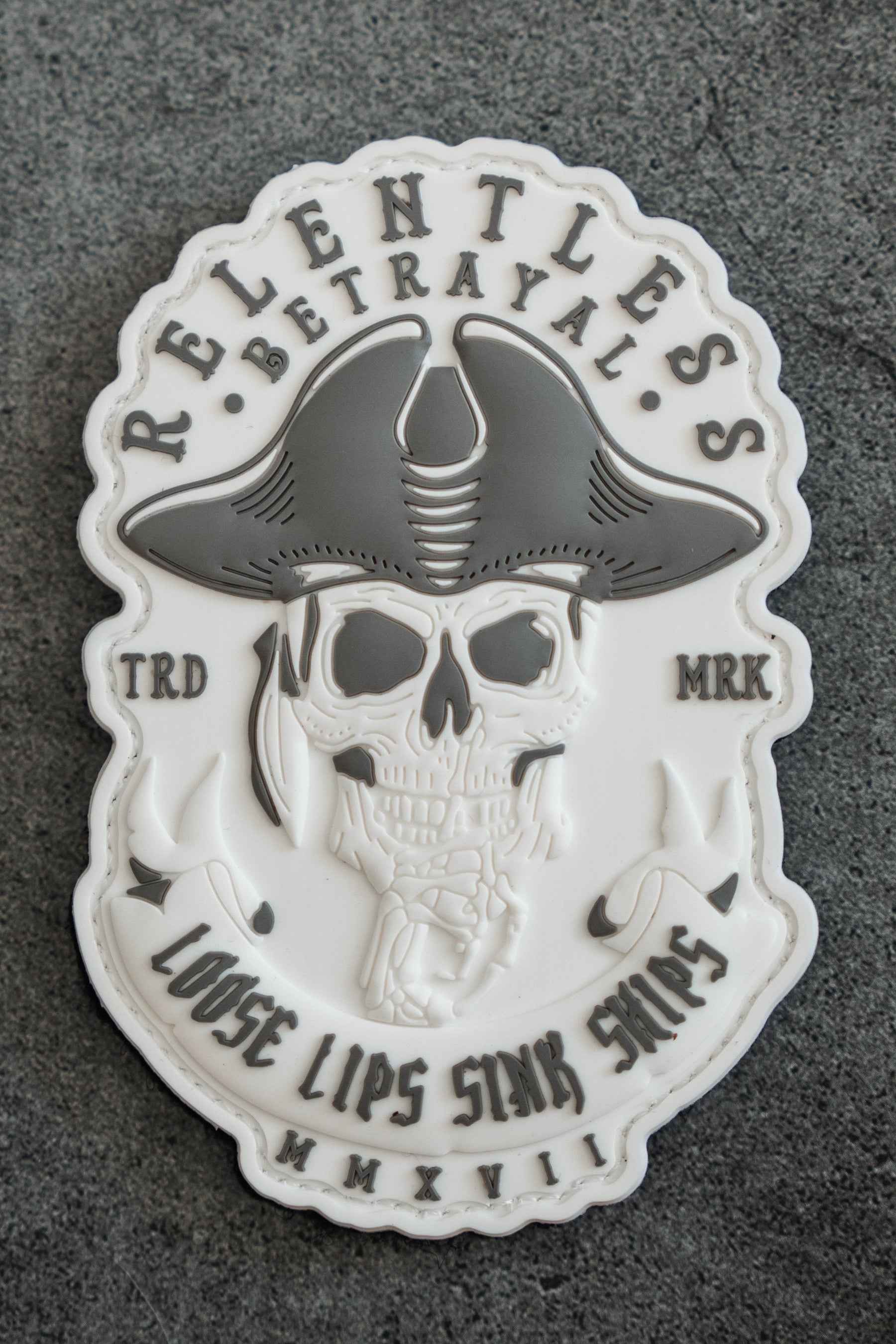 An embossed white sticker becomes a 3D PVC patch featuring a pirate skull with a tricorn hat. It has a textured gray background with Relentless Betrayal, Loose Lips Sink Ships, and MMXVII in gray text, reminiscent of WWIIs grit, by Relentless Betrayal.