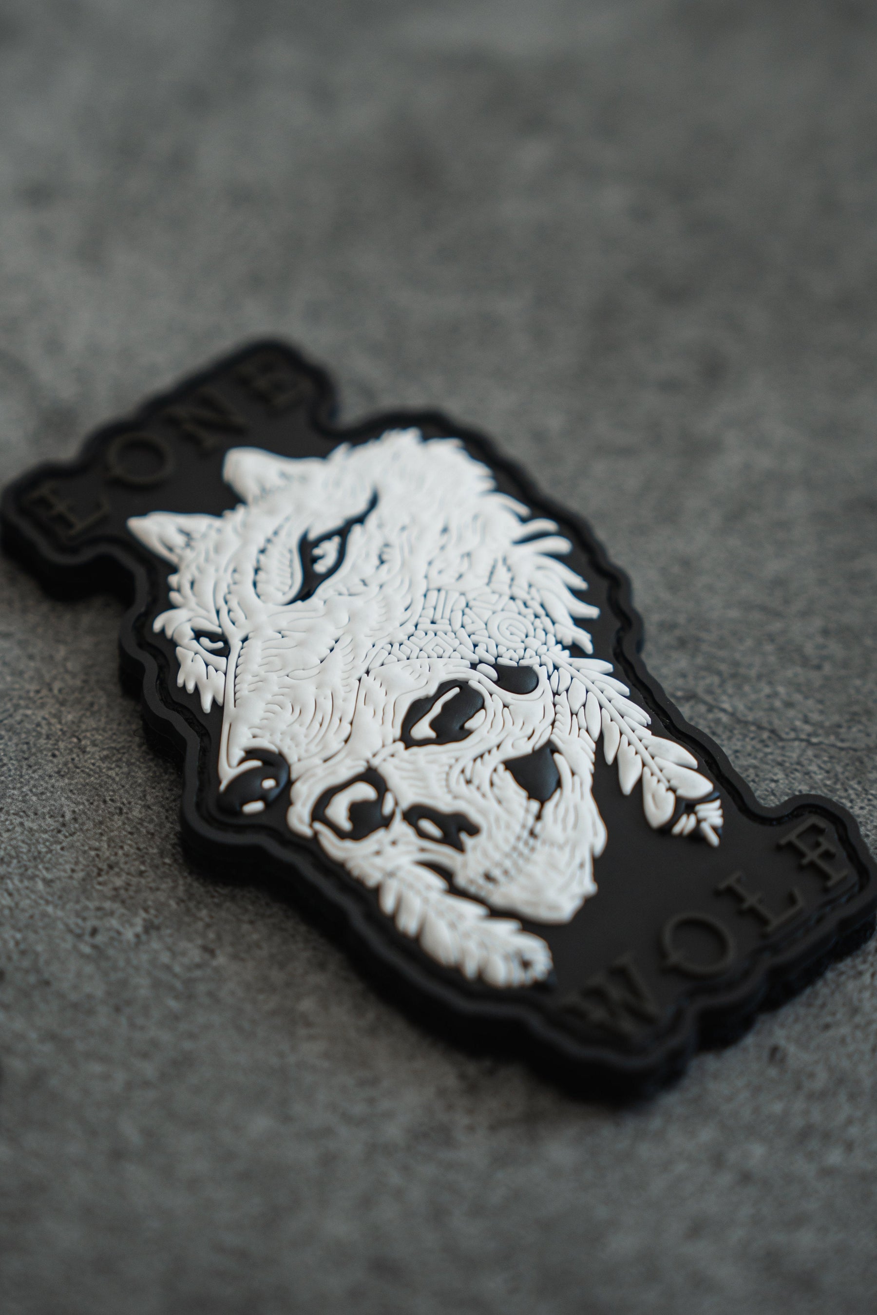 The Lone Wolf PVC Patch by Relentless Betrayal showcases a detailed wolf design in black and white, with LONE WOLF boldly on either side. It features Velcro backing for easy attachment and rests on a textured gray surface, adding mythical allure to its rugged charm.
