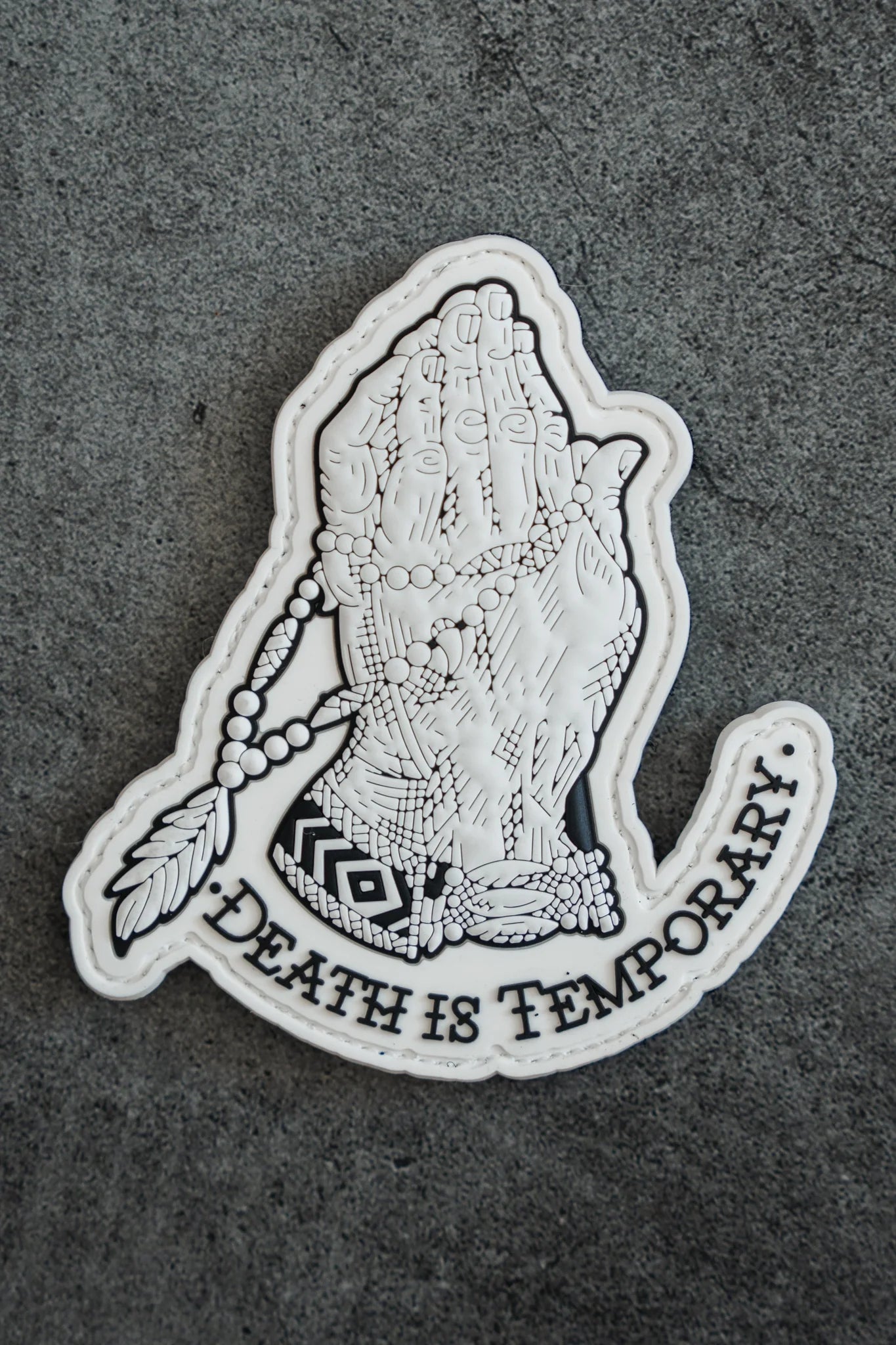 The Death Is Temporary PVC Patch by Relentless Betrayal showcases a 3D decal of two praying hands with beads and a feather, set on a textured surface, featuring the message Death is Temporary to symbolize renewal and transformation.
