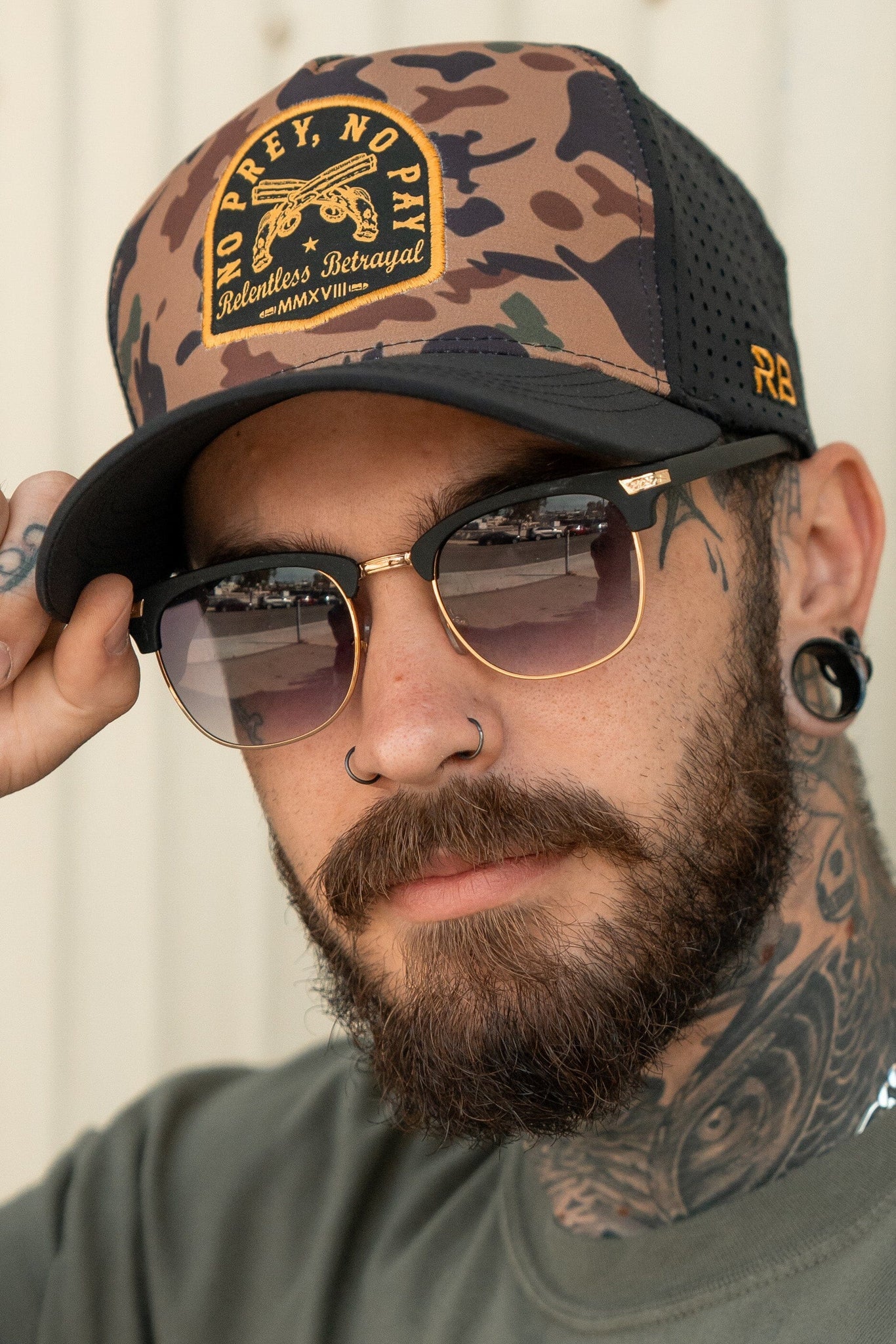 A bearded man with sunglasses, a nose piercing, and neck tattoos sports a Relentless Betrayal No Prey, No Pay Frog Camo Hat. His edgy look is completed by visible ear gauges as he adjusts his shades, embracing the pirate lifestyle with intent while gazing at the camera.