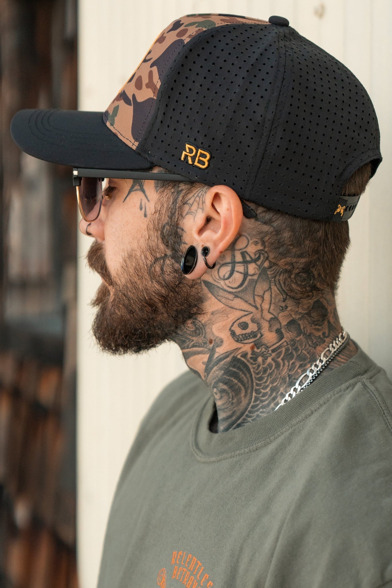 A bearded man with neck tattoos and ear gauges wears sunglasses and a black and camo No Prey, No Pay Frog Camo Hat by Relentless Betrayal, featuring RB on the side. Embracing the pirate lifestyle, he finishes his look with a green shirt and a silver chain.