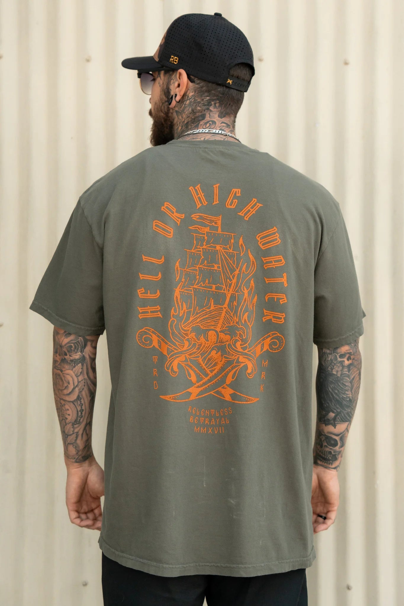 A man in a black cap and sunglasses faces away, showcasing the Hell or High Water Premium Tee - Olive by Relentless Betrayal, featuring an orange fiery ship symbolizing resilience. His full sleeve tattoos and unwavering spirit stand tall against the corrugated wall.