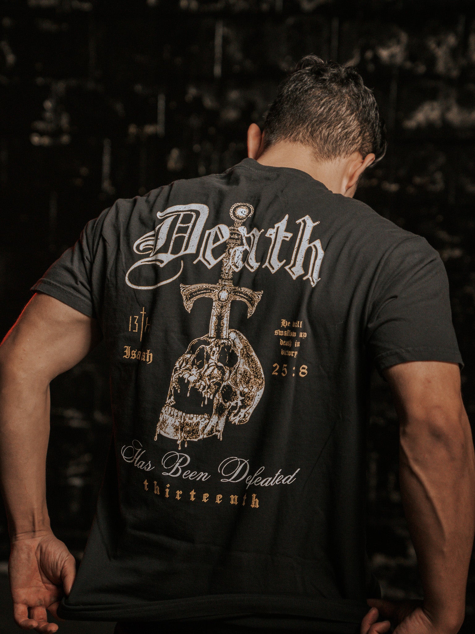 A man, facing away from the camera, wears the Deaths Defeat Premium Tee by 13th. The black shirt showcases a skull and sword with Death has been defeated plus references like Isaiah 25:8. The dark, textured backdrop symbolizes victory over death.