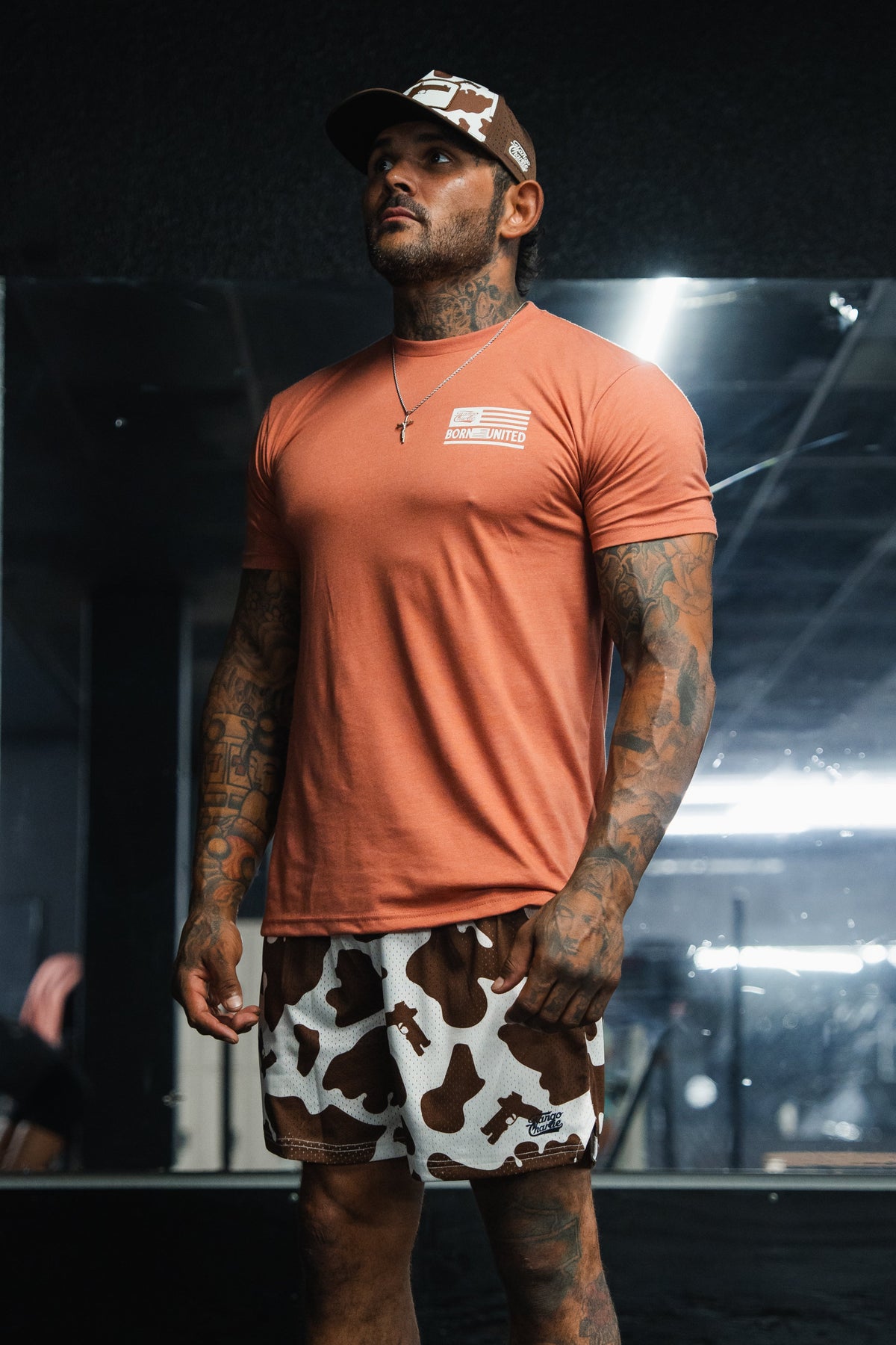 A man wears a coral Tango Charlie Apparel LHSG Moo-Moo Pew Tee, brown and white cow-print athletic shorts, and a matching cow-print baseball cap.
