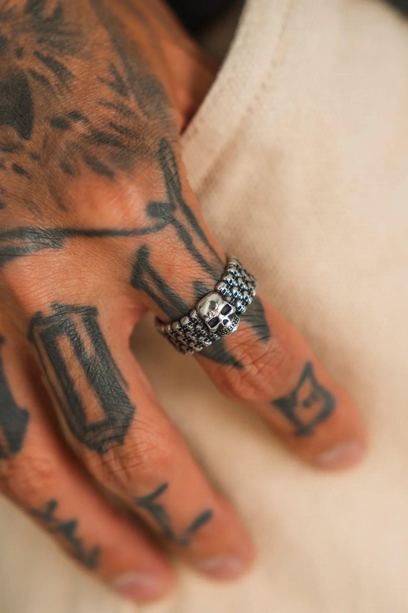 A tattooed hand rests on fabric, wearing the Relentless Betrayal Kill Switch Ring—a handcrafted stainless steel ring featuring a skull design and textured band—on the middle finger.