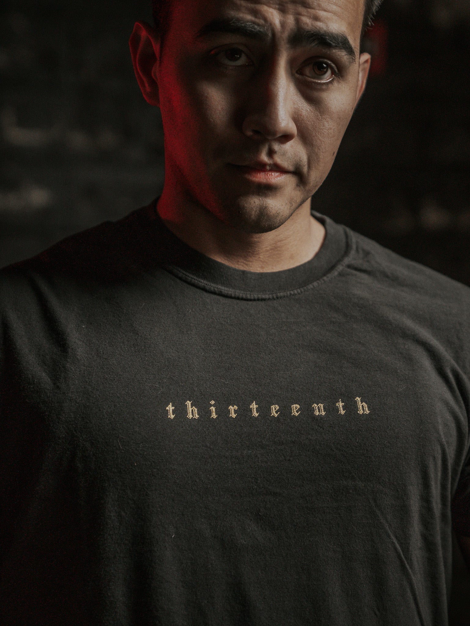 A person wears the Deaths Defeat Premium Tee by 13th, featuring light-colored thirteenth text on a dark shirt. In the dim red-tinged background, theres a subtle echo of victory over darkness, reminiscent of Isaiah 25:8.