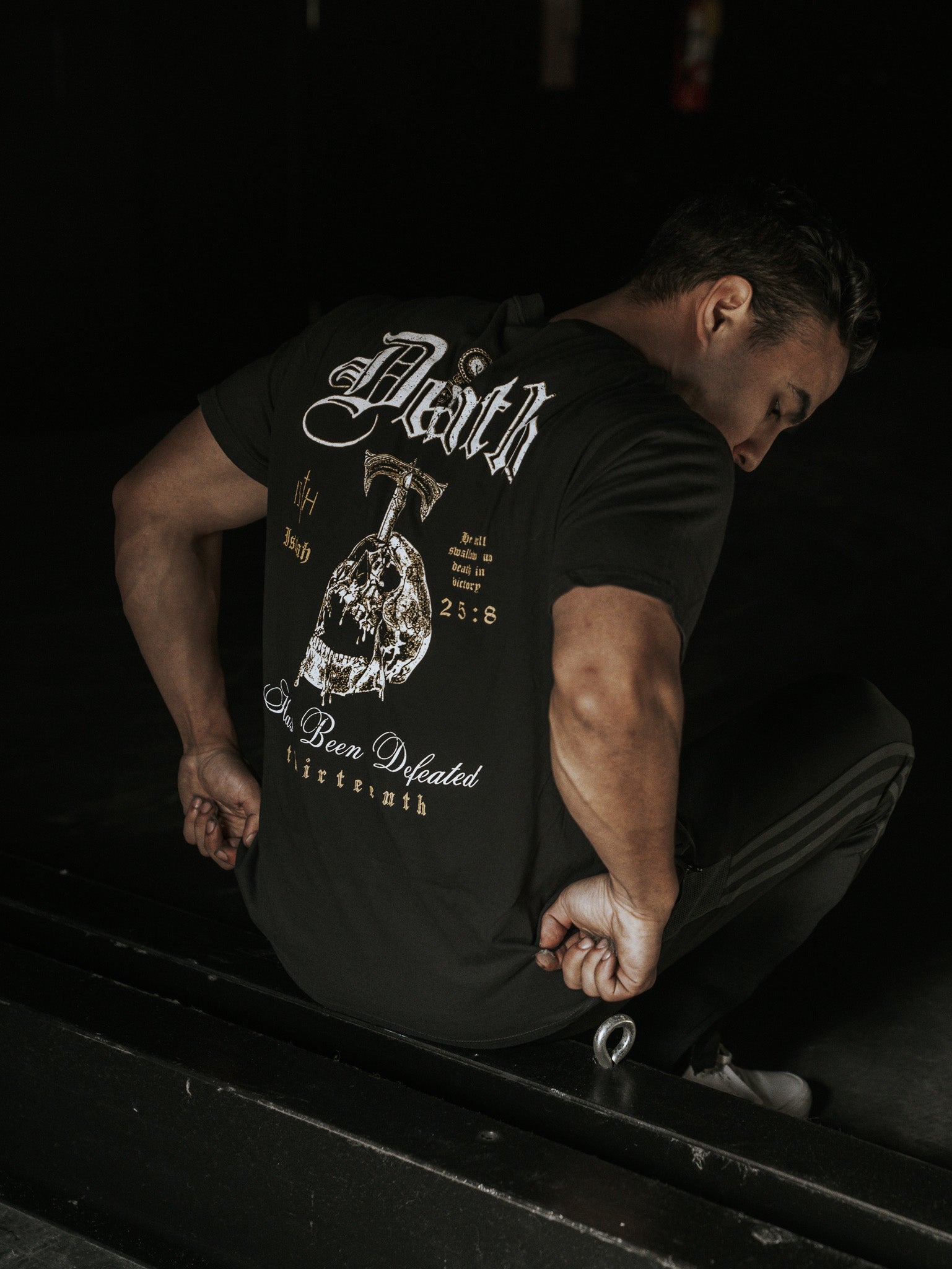 A man crouches on a dark floor, facing away, wearing the Deaths Defeat Premium Tee by 13th. The black shirt features bold text and graphic designs, including a cross and Faith, as he leans forward. The dark background evokes triumph over death as reflected in Isaiah 25:8.