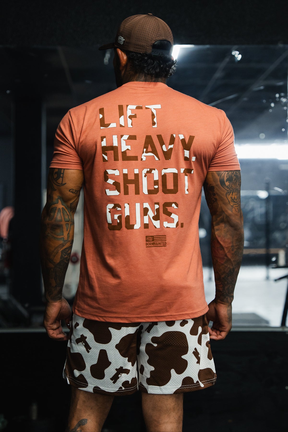 A person seen from behind wears an orange LHSG Moo-Moo Pew Tee with brown camo lettering and cow-print athletic shorts.