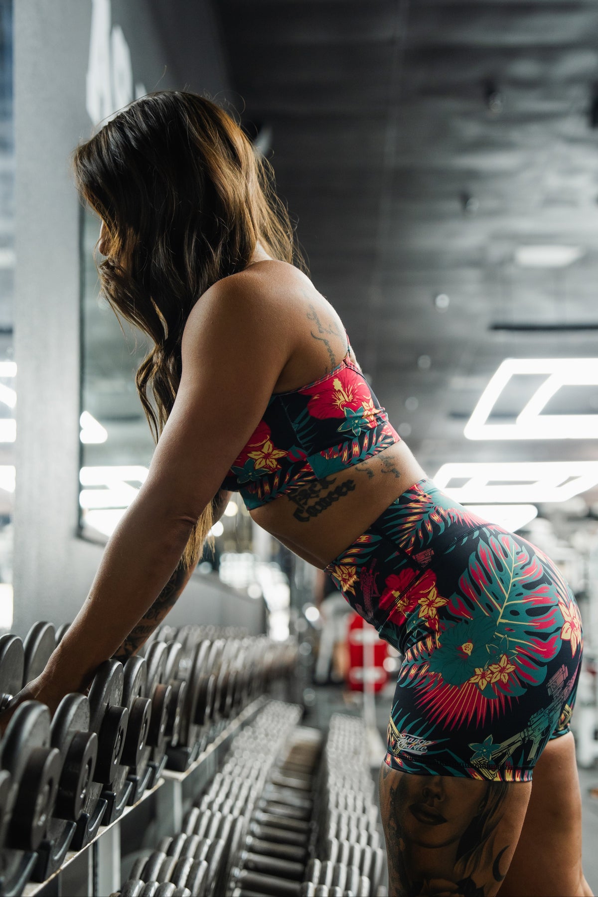 A woman in Tango Charlie Apparel's Women's High-Waisted Seamless Front Compression Training Shorts | Tropical Assassin leans over a rack of dumbbells in a modern, brightly lit gym, preparing to lift weights.