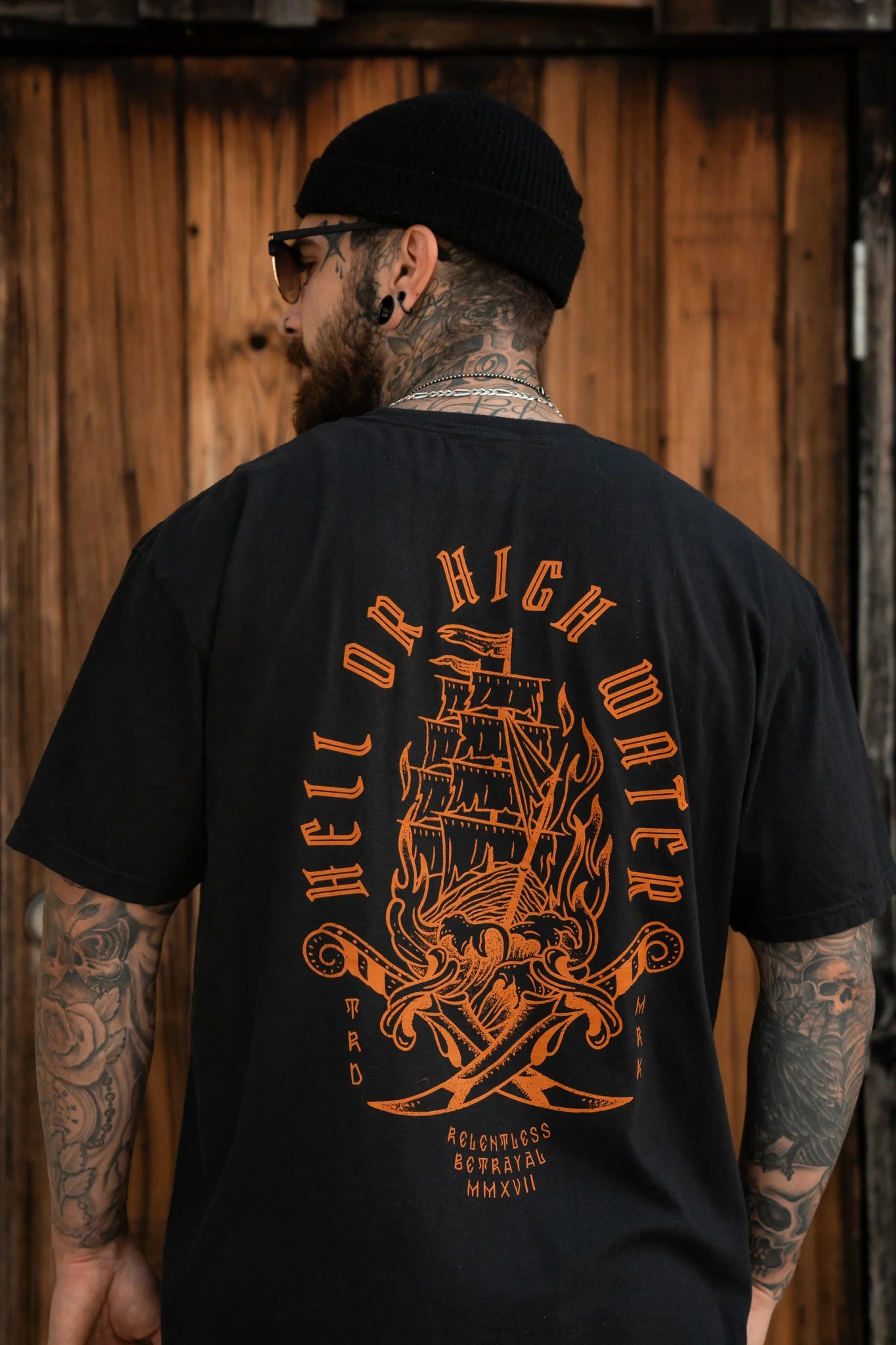 A tattooed man wearing a beanie and the Hell or High Water Premium Tee by Relentless Betrayal, showcasing an orange ship design on a black T-shirt, exudes unwavering spirit. The backdrop is framed with wooden panels.