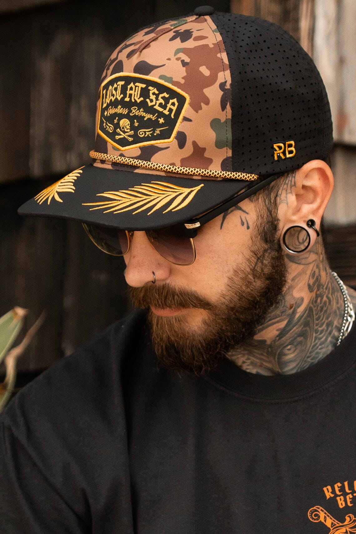 A bearded, tattooed man embodies rugged adventure wearing Relentless Betrayals Lost At Sea Frog Camo Snapback. With glasses and large earrings, he sits outdoors against wood, dressed in black.