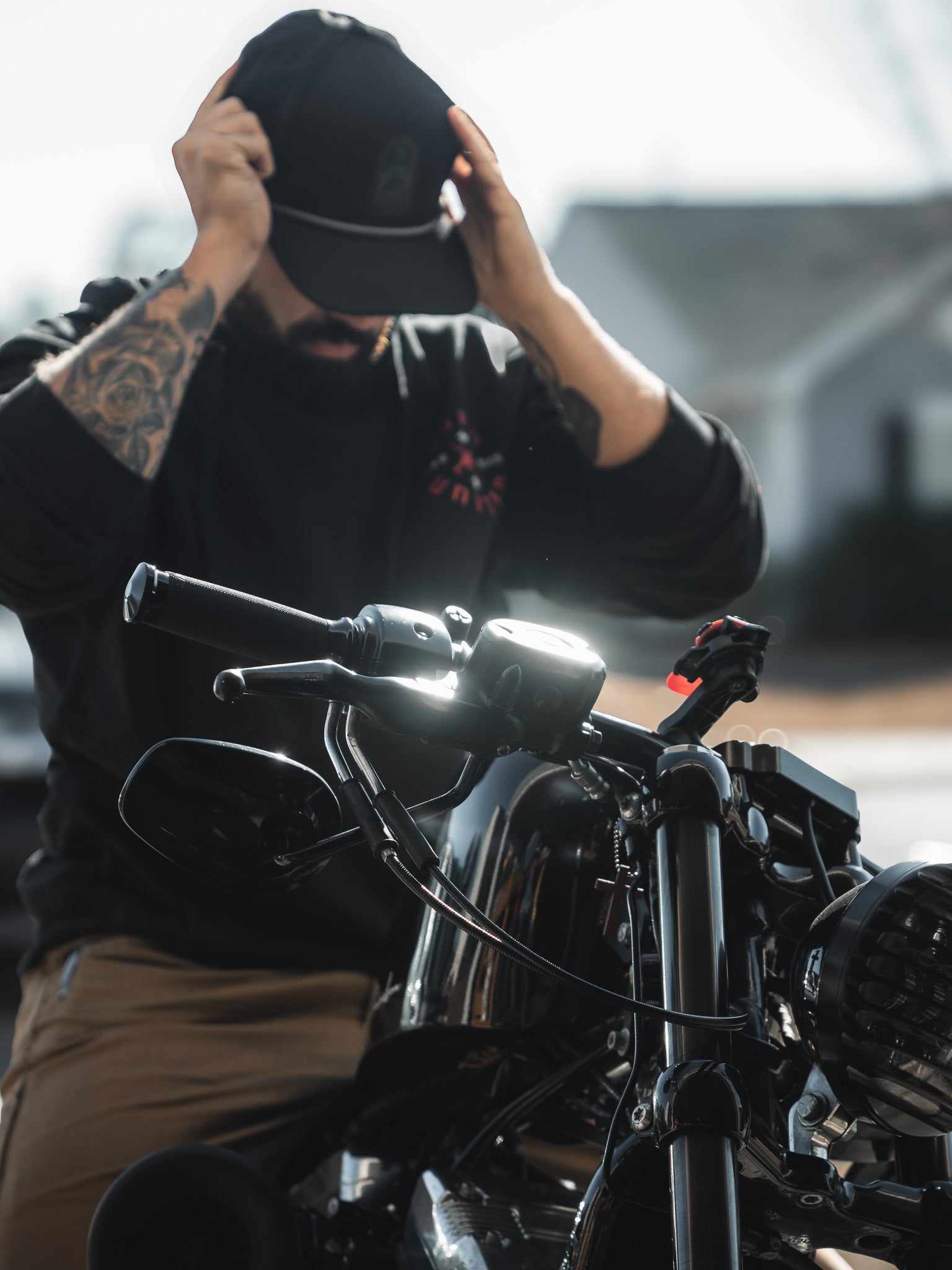 A tattooed man, embodying the spirit of a modern-day saint, adjusts his Saints and Sinners Hat by Born United Private Label while sitting on a motorcycle. The bikes handlebars are in focus as sunlight casts reflections on its glossy surface.