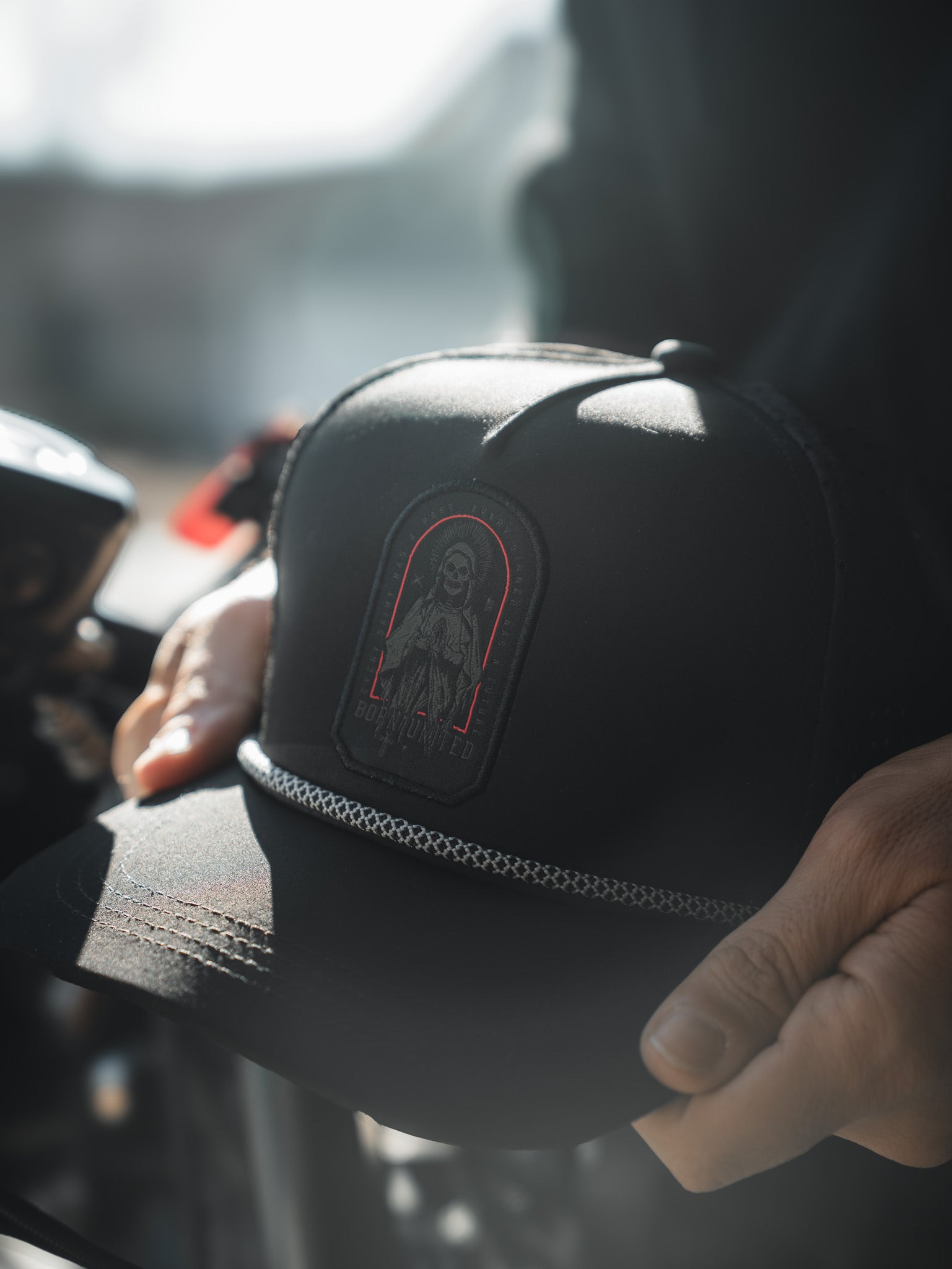 A person holds the Saints and Sinners Hat by Born United Private Label, a black cap with an embroidered cloaked figure, red outline, and decorative front chain. Soft lighting highlights this mysterious piece.
