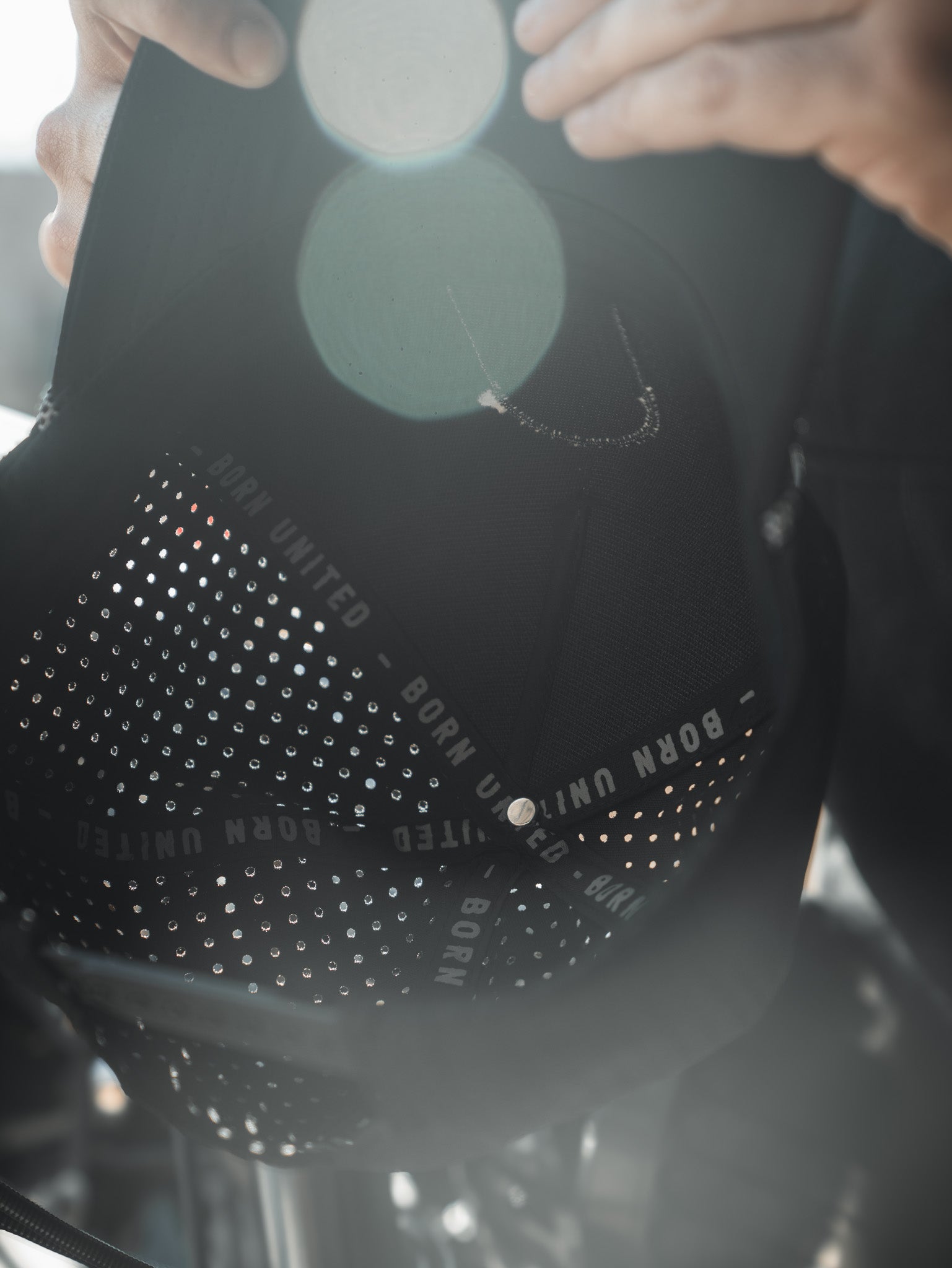 A close-up image shows the Saints and Sinners Hat from Born United Private Label, featuring BORN UNITED in white on the inner band. The black cap is dotted with small perforations, accentuated by lens flares. A partially visible person holds it reverently, cherishing the sense of unity it represents.