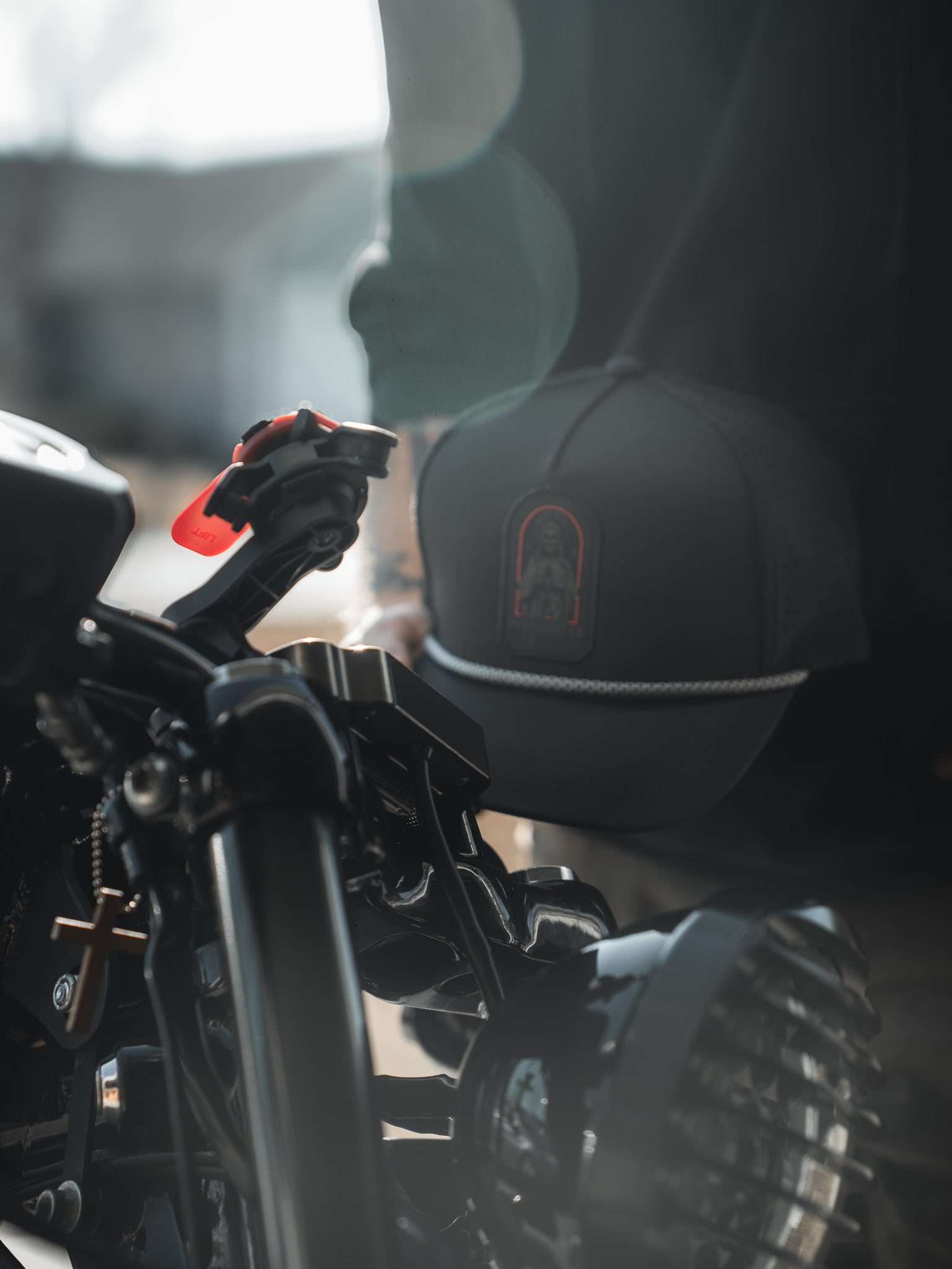 A close-up shows a motorcycle handlebar and headlight, with a person wearing the Born United Private Label Saints and Sinners Hat. The subtle saintly design on the hat catches the sunlight, creating an ethereal lens flare effect in the background.