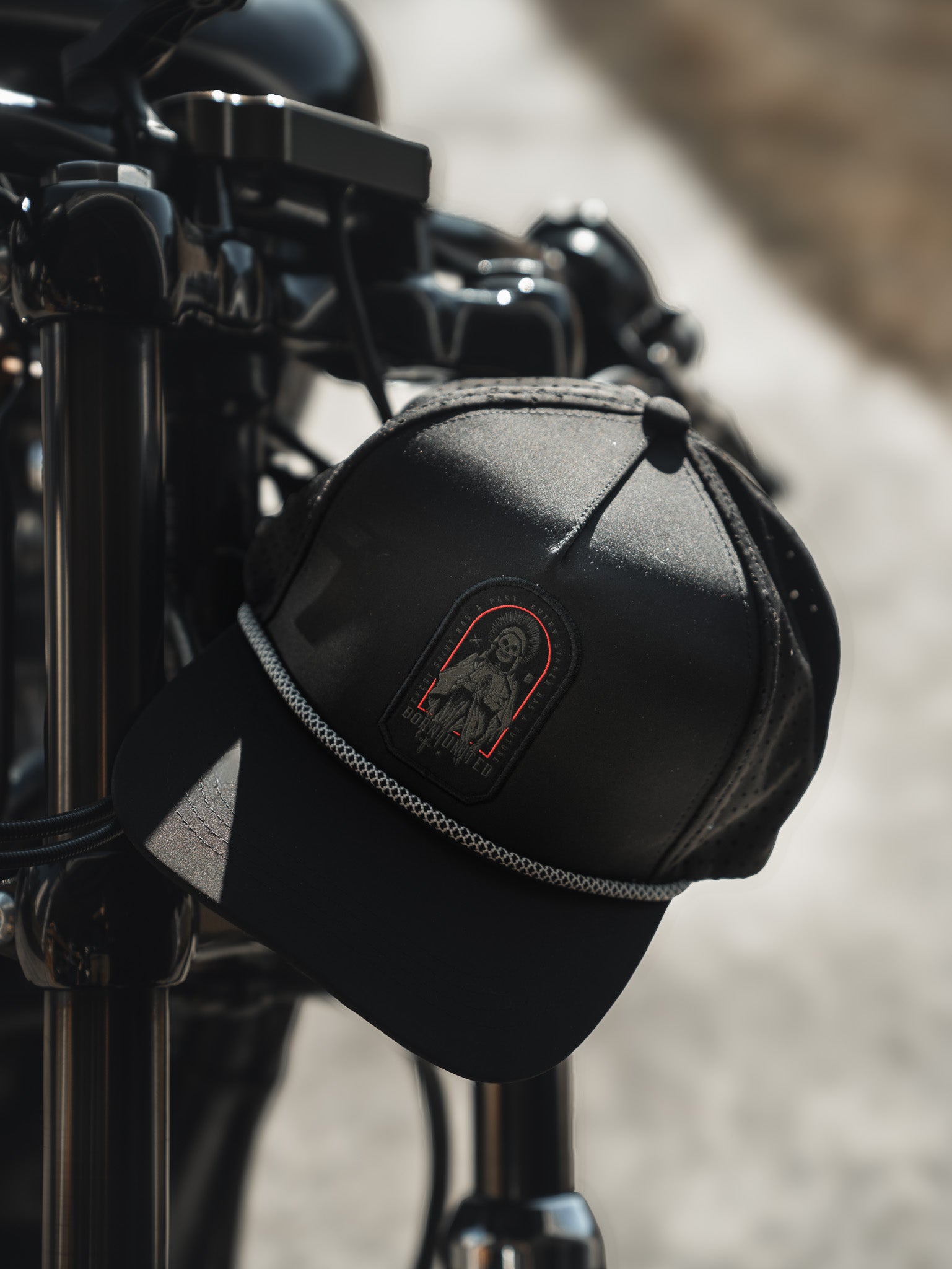 A Saints and Sinners Hat from Born United Private Label, featuring a Saint or Sinner? patch with a red outline of a woman, hangs on a motorcycle handlebar. Its textured band adds flair against the blurred outdoor setting, hinting at an adventure awaiting its owner.