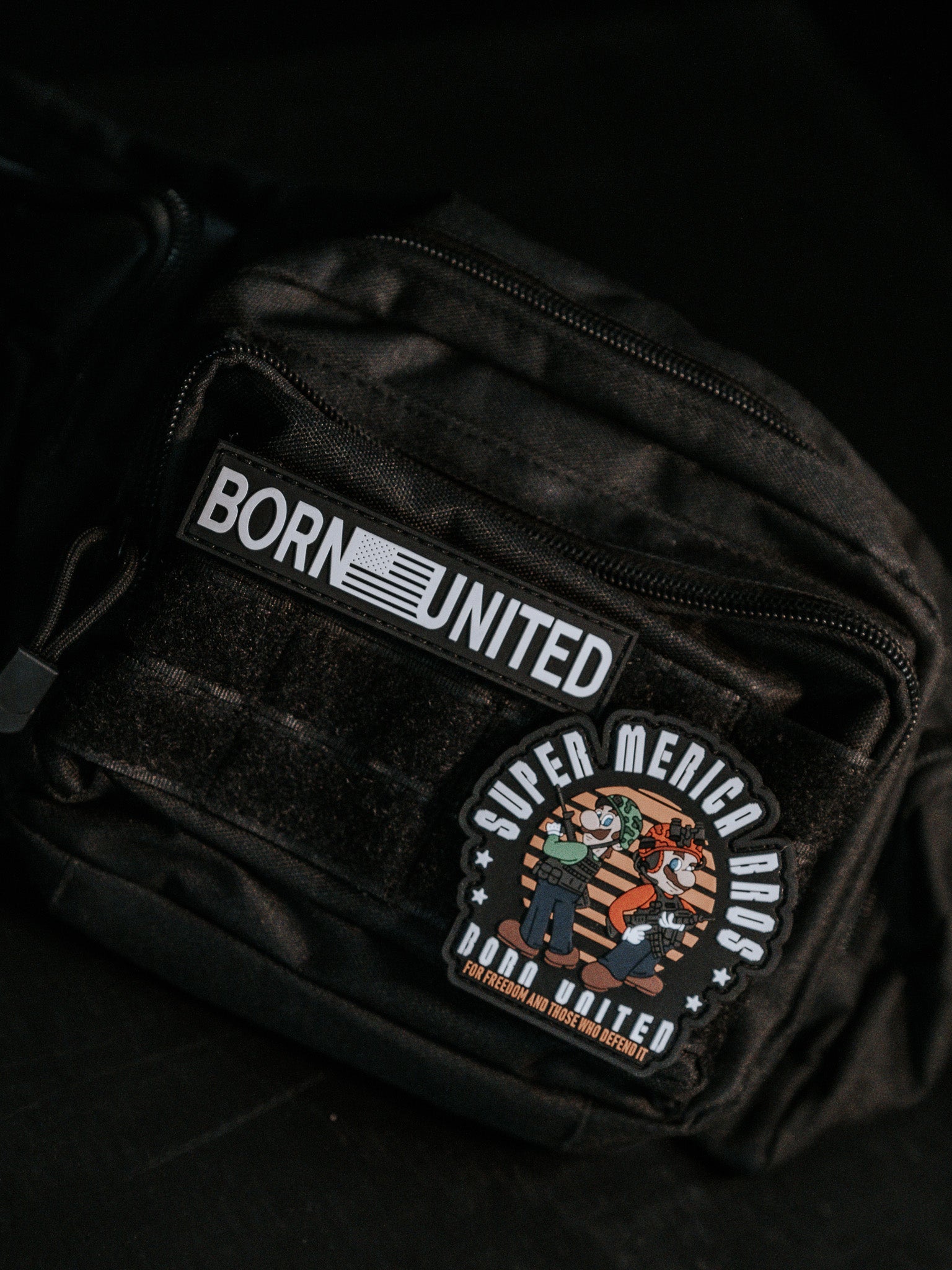 The Midnight Black Tac-Fanny-Pac by Born United Private Label features a BORN UNITED patch and a SUPER MERICA BROS. round patch with playful hero characters, adding a whimsical touch to its sleek design.