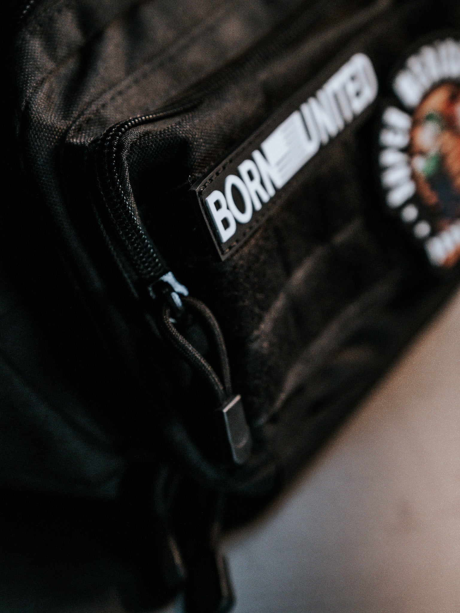 Close-up of a black Midnight Black Tac-Fanny-Pac by Born United Private Label. A patch with BORN UNITED in white letters is seen on a zippered pocket, revealing the fabric texture, while another colorful patch is partially visible.