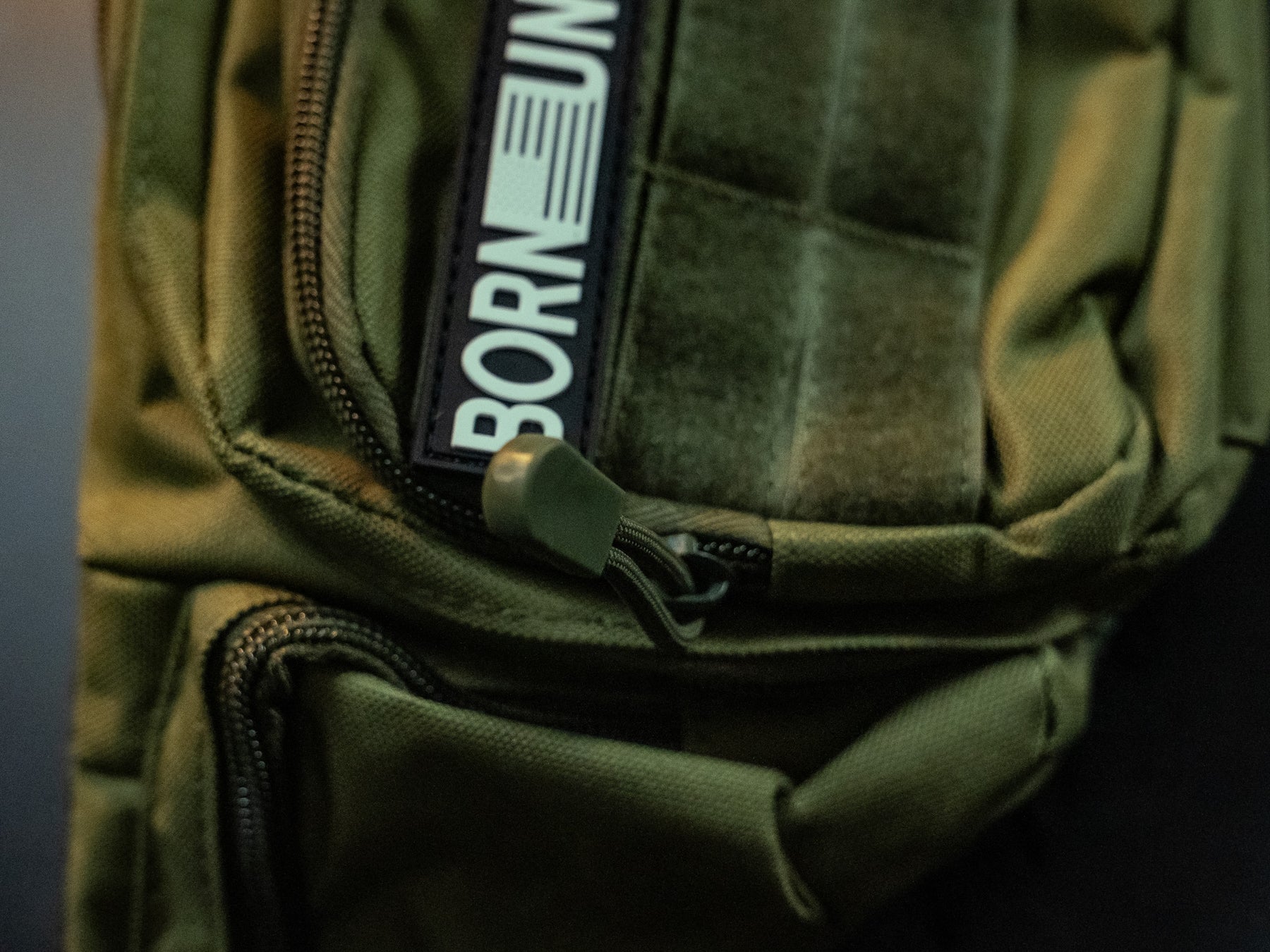 A close-up of the OD Green Tac-Fanny-Pac by Born United Private Label shows a visible zipper and patch with BORN, followed by an unreadable section. Dim lighting highlights the fabric texture and zipper pull, enhancing its rugged style.