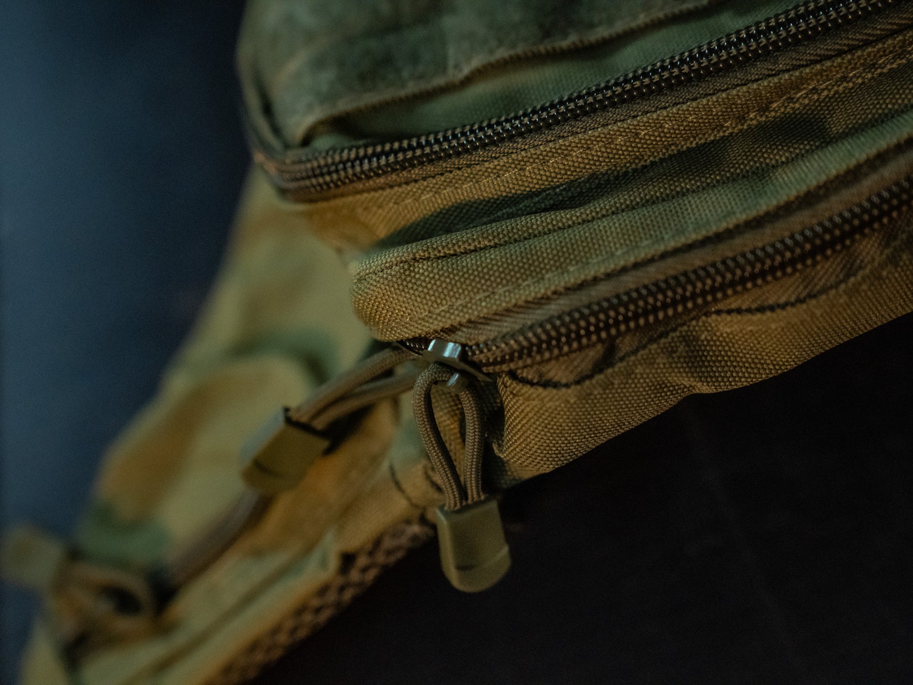 The OD Green Tac-Fanny-Pac by Born United Private Label is shown in a close-up, highlighting its tactical canvas texture, stitching, and durable zipper pulls. The low-light setting emphasizes its rugged design, resembling an oversized fanny pack.