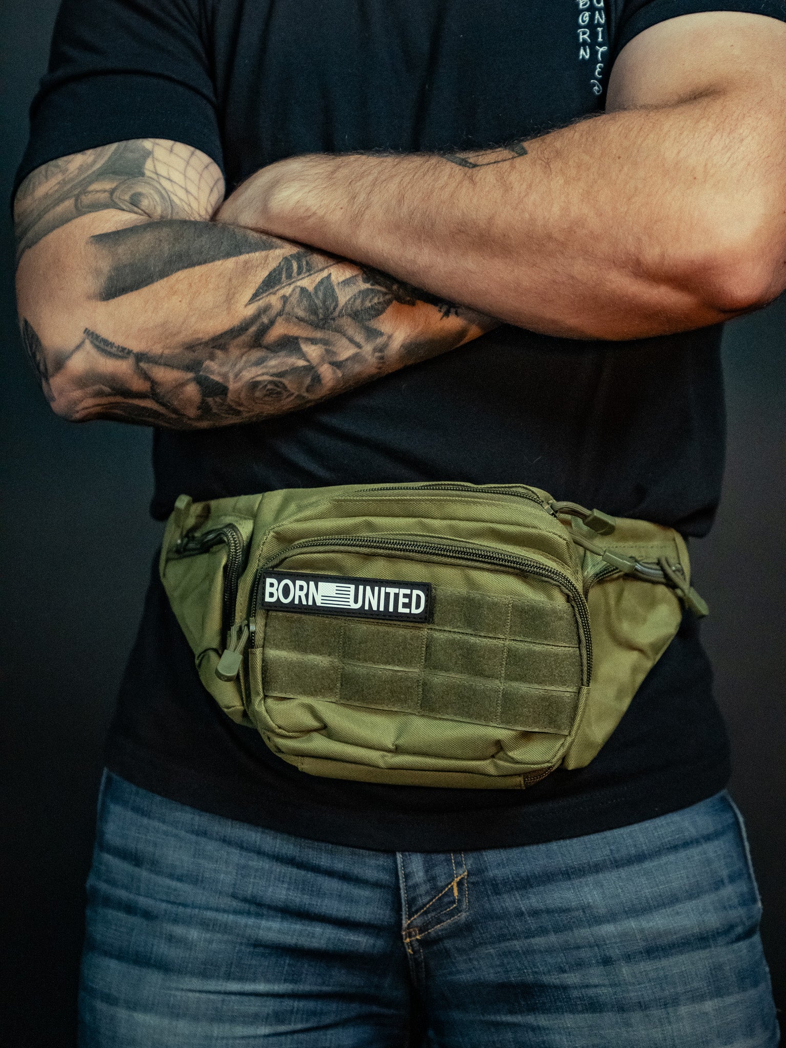 A person with crossed arms, wearing a black shirt and blue jeans, sports an OD Green Tac-Fanny-Pac by Born United Private Label. Their left arm is adorned with a floral tattoo against a dark background.
