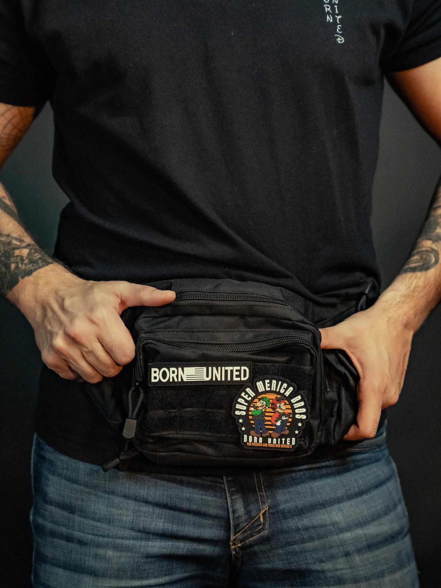 Clad in a black T-shirt and jeans, a person holds the Midnight Black Tac-Fanny-Pac from Born United Private Label, featuring patches like BORN UNITED and vibrant graphics. Their forearms display tattoos that pop against the dark backdrop.