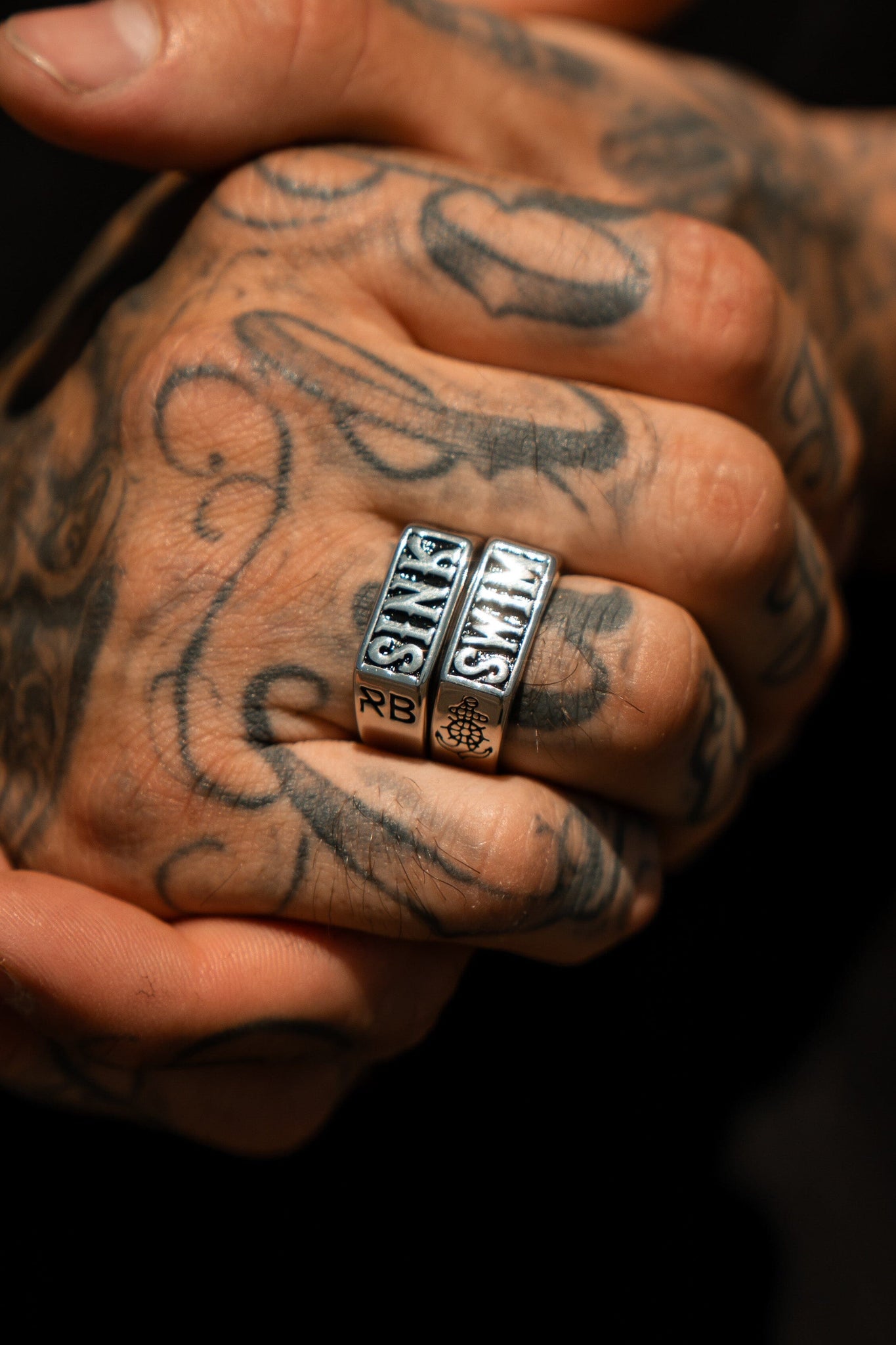 Close-up of a tattooed hand wearing two handcrafted Sink or Swim Rings by Relentless Betrayal on the middle and ring fingers, each engraved with SANE. The intricate tattoos feature swirls and patterns covering the knuckles and fingers.