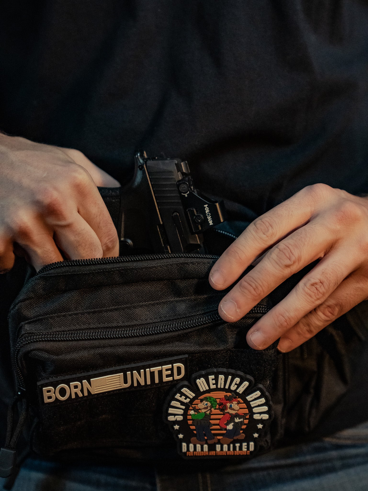 A person in a black shirt discreetly tucks a handgun into the Midnight Black Tac-Fanny-Pac by Born United Private Label, featuring two patches: one with BORN UNITED and another showcasing a colorful SUPER MERICA RAC design.
