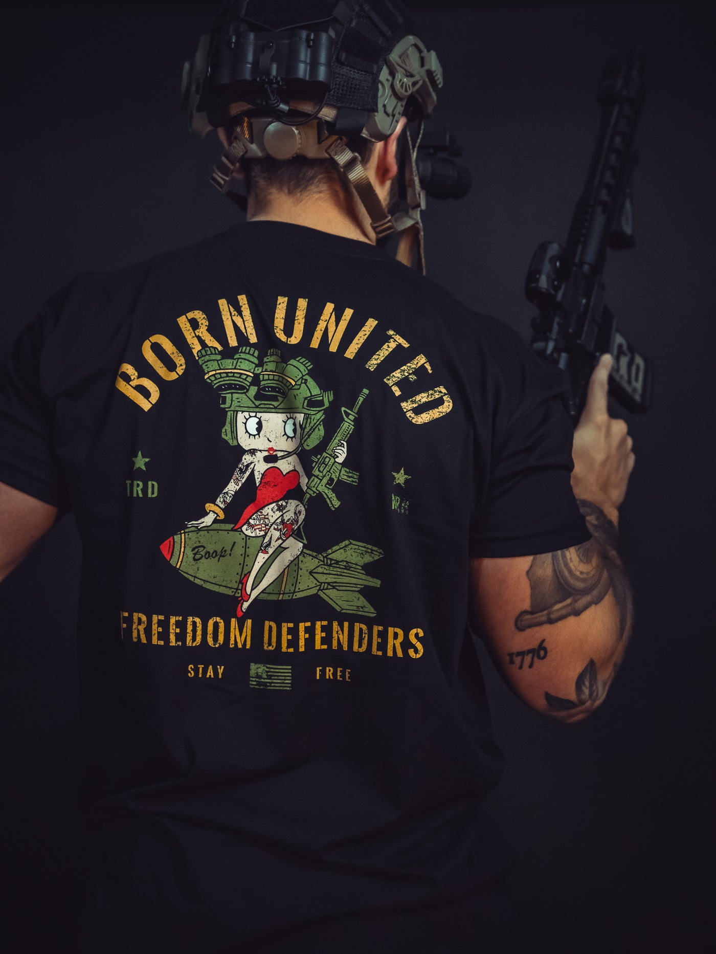Seen from behind, a person in tactical gear holds a rifle, wearing the Betty Boop Tee by Born United Private Label. The T-shirt features an illustrated character sitting on a bomb alongside the bold text: Born United, Freedom Defenders, Stay Free, symbolizing their dedication to freedom.