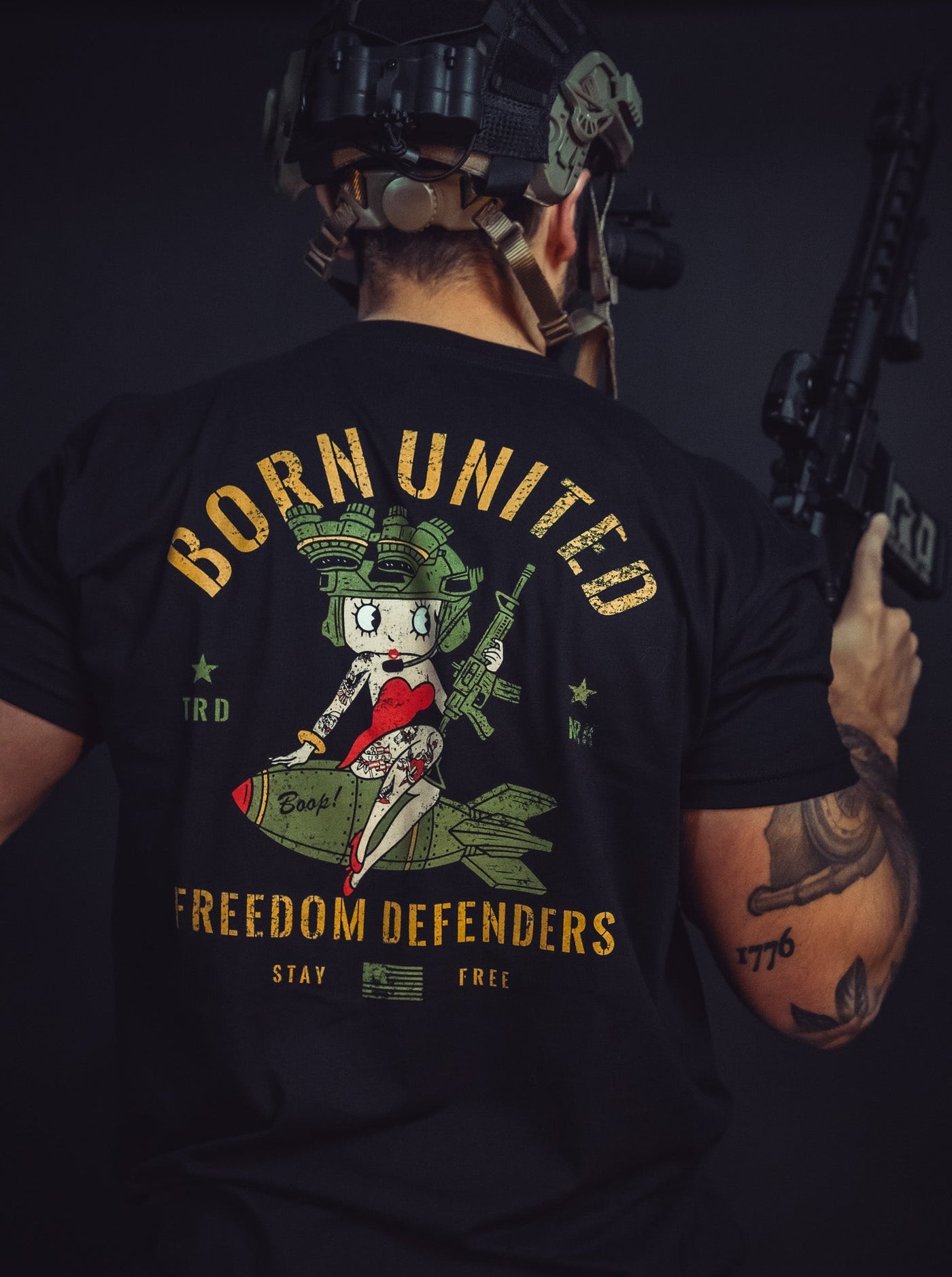 Seen from behind, a person in tactical gear holds a rifle, wearing the Betty Boop Tee by Born United Private Label. The T-shirt features an illustrated character sitting on a bomb alongside the bold text: Born United, Freedom Defenders, Stay Free, symbolizing their dedication to freedom.