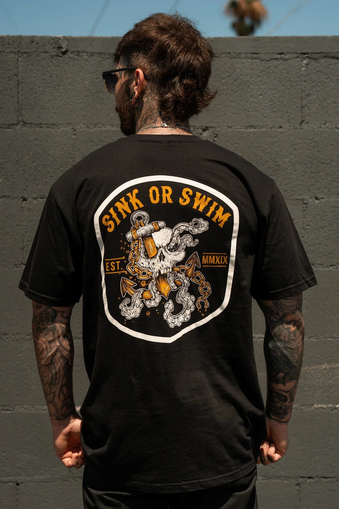 A tattooed individual faces a gray brick wall wearing the Relentless Betrayal Sink Or Swim Kraken T-Shirt, which showcases a graphic octopus with chains and anchor plus "SINK OR SWIM EST. MMXIX" text on the back.
